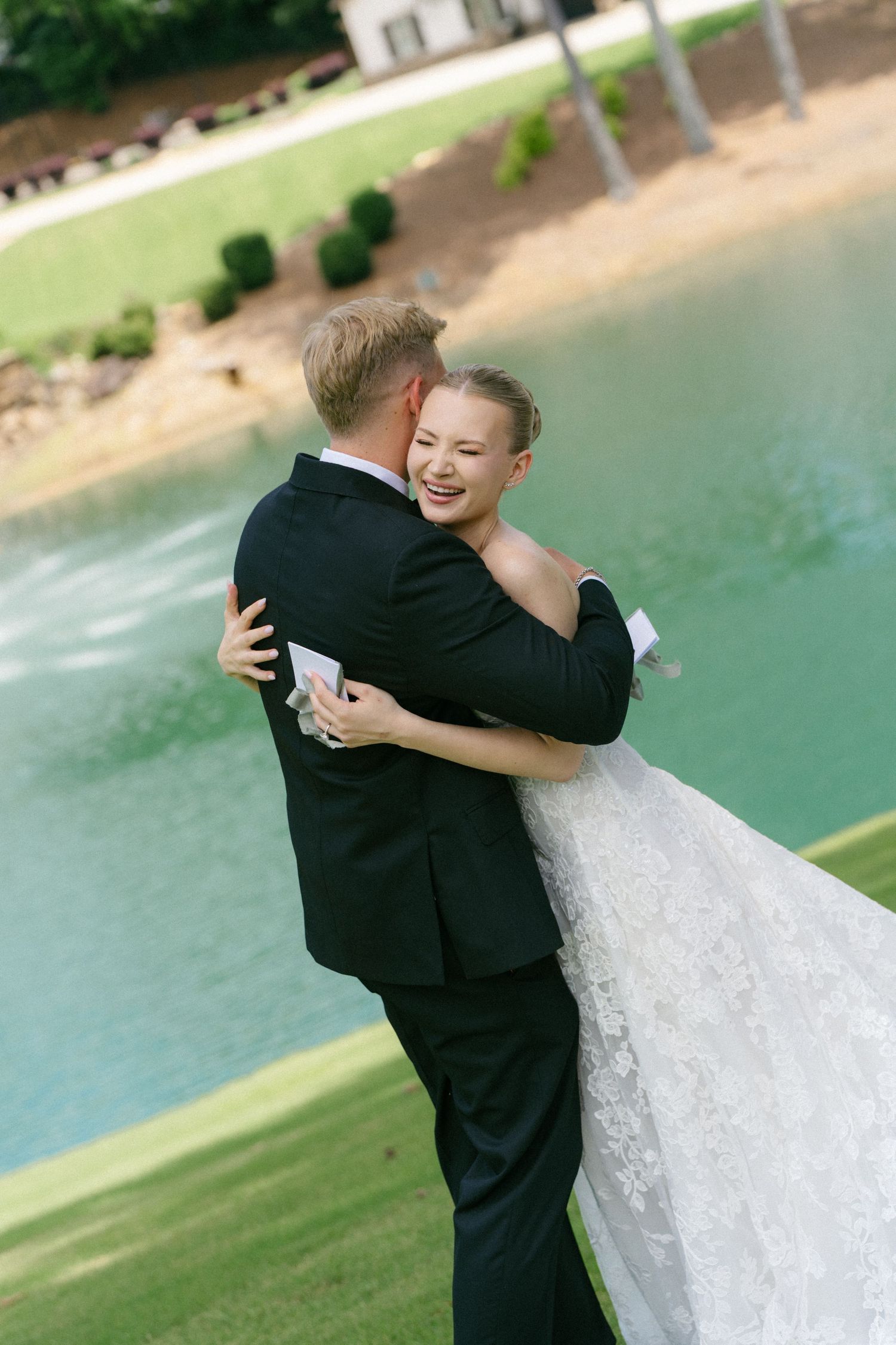 Joyful bride in lace gown embraces groom in black suit outdoors near a turquoise pond on their wedding day.
