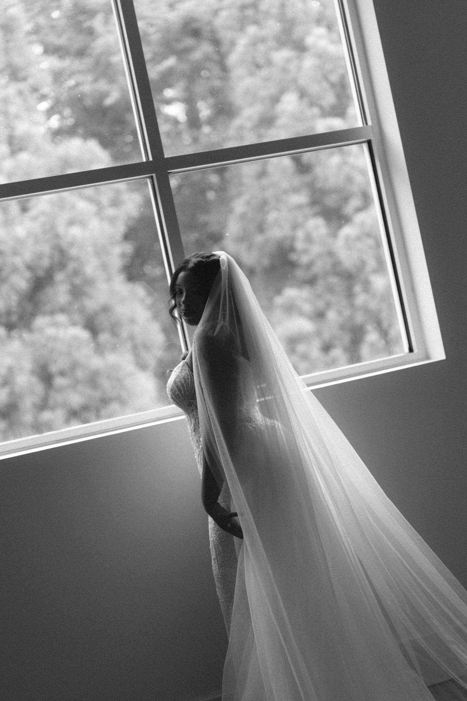 Bride in lace gown and veil silhouetted against large window, black and white photo.