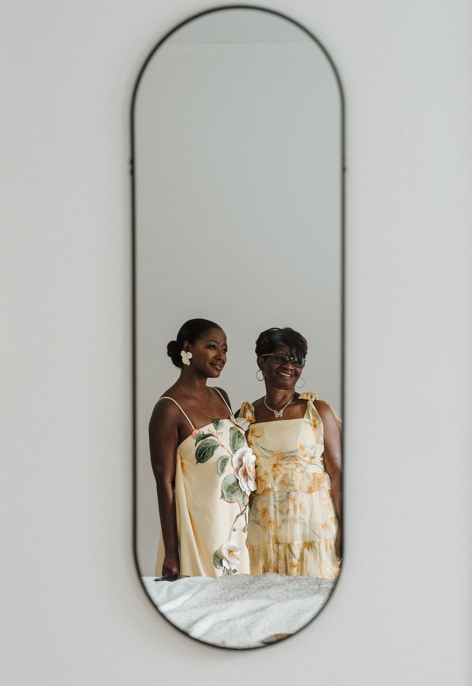 Two women in elegant yellow dresses smiling together, reflected in an oval mirror on a white wall.