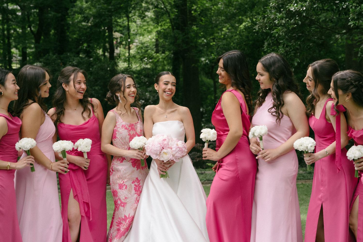 Bride in white gown with bridesmaids in pink dresses holding bouquets, laughing outdoors with green trees background.