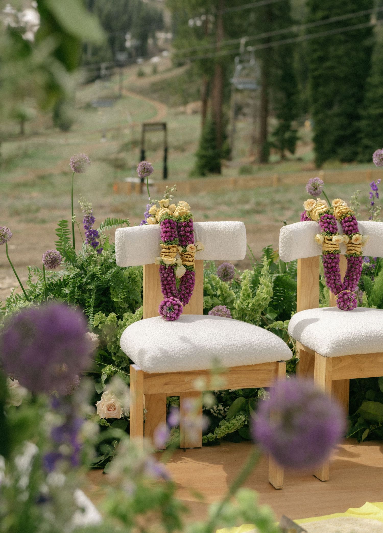 Wooden ceremony chairs adorned with purple floral garlands and greenery, set against a scenic mountain backdrop.