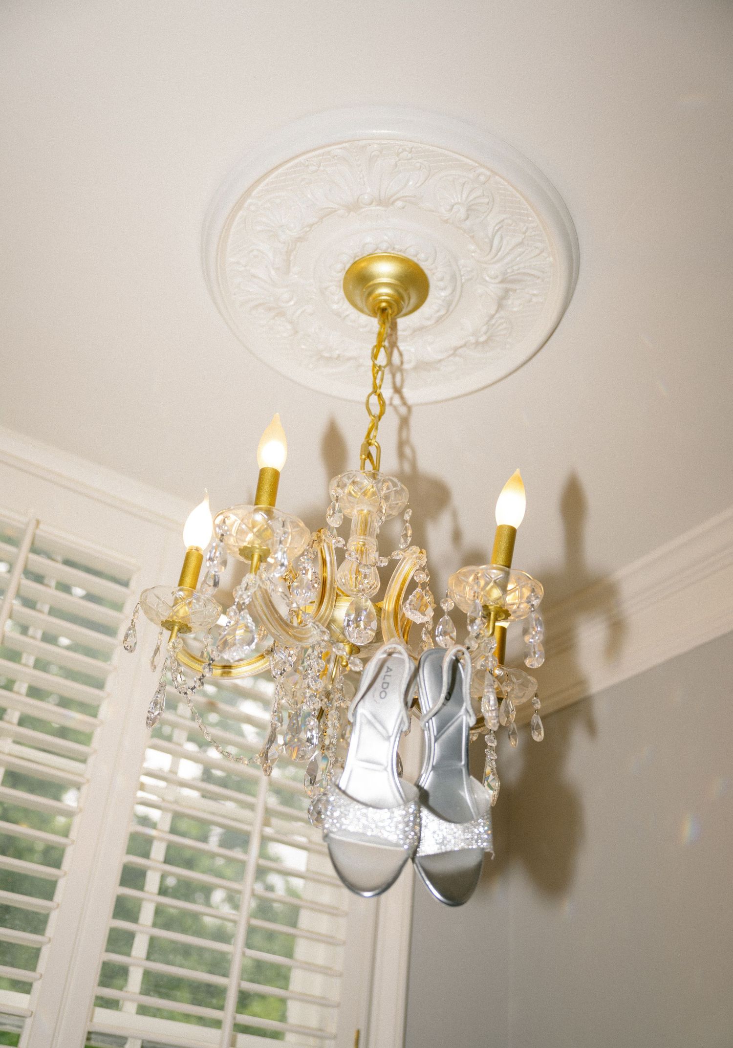 Silver bridal heels hanging from a gold crystal chandelier with candle lights mounted on white ceiling.