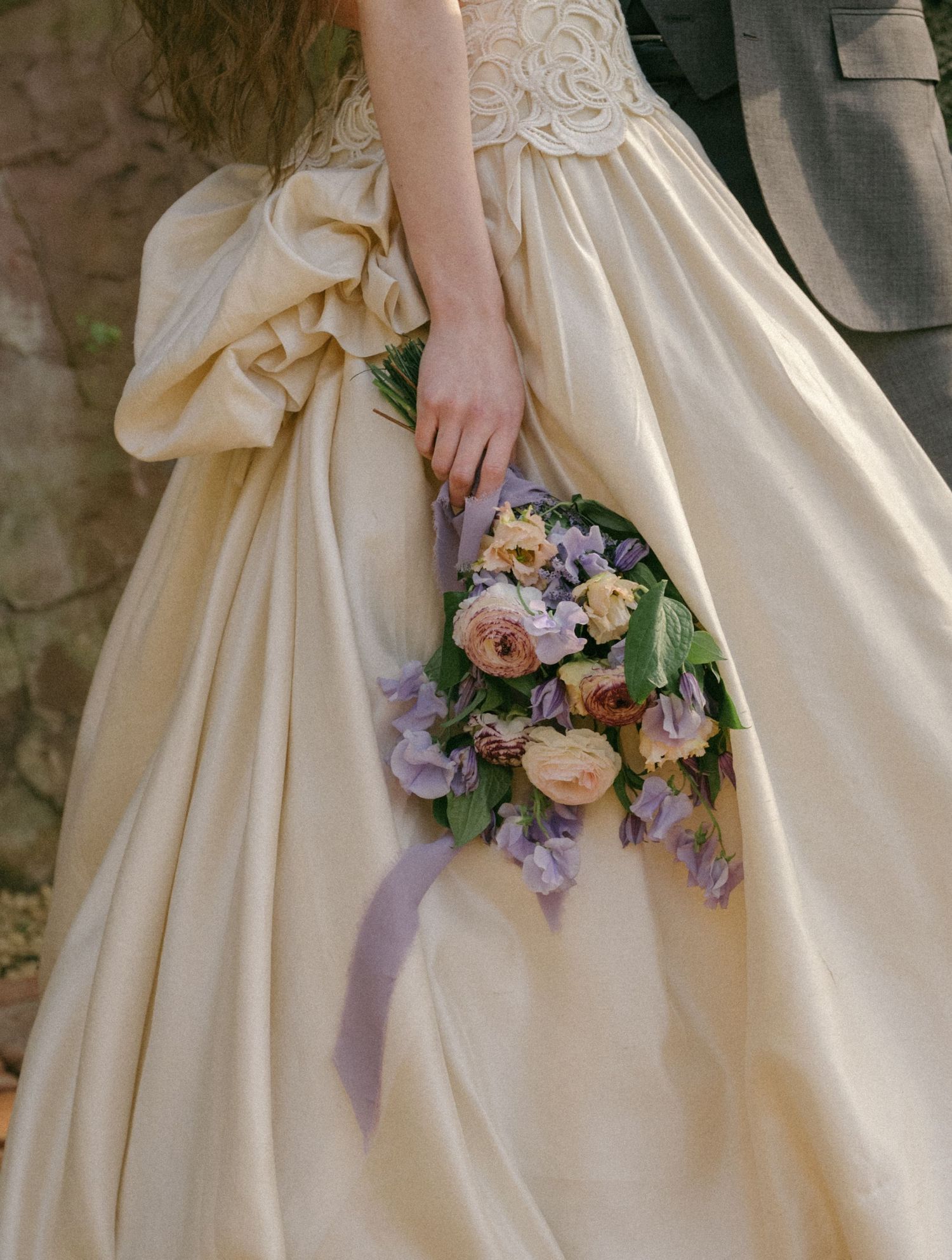 Bride in ivory ballgown holding purple and pink floral bouquet with ribbon, standing beside groom in gray suit.