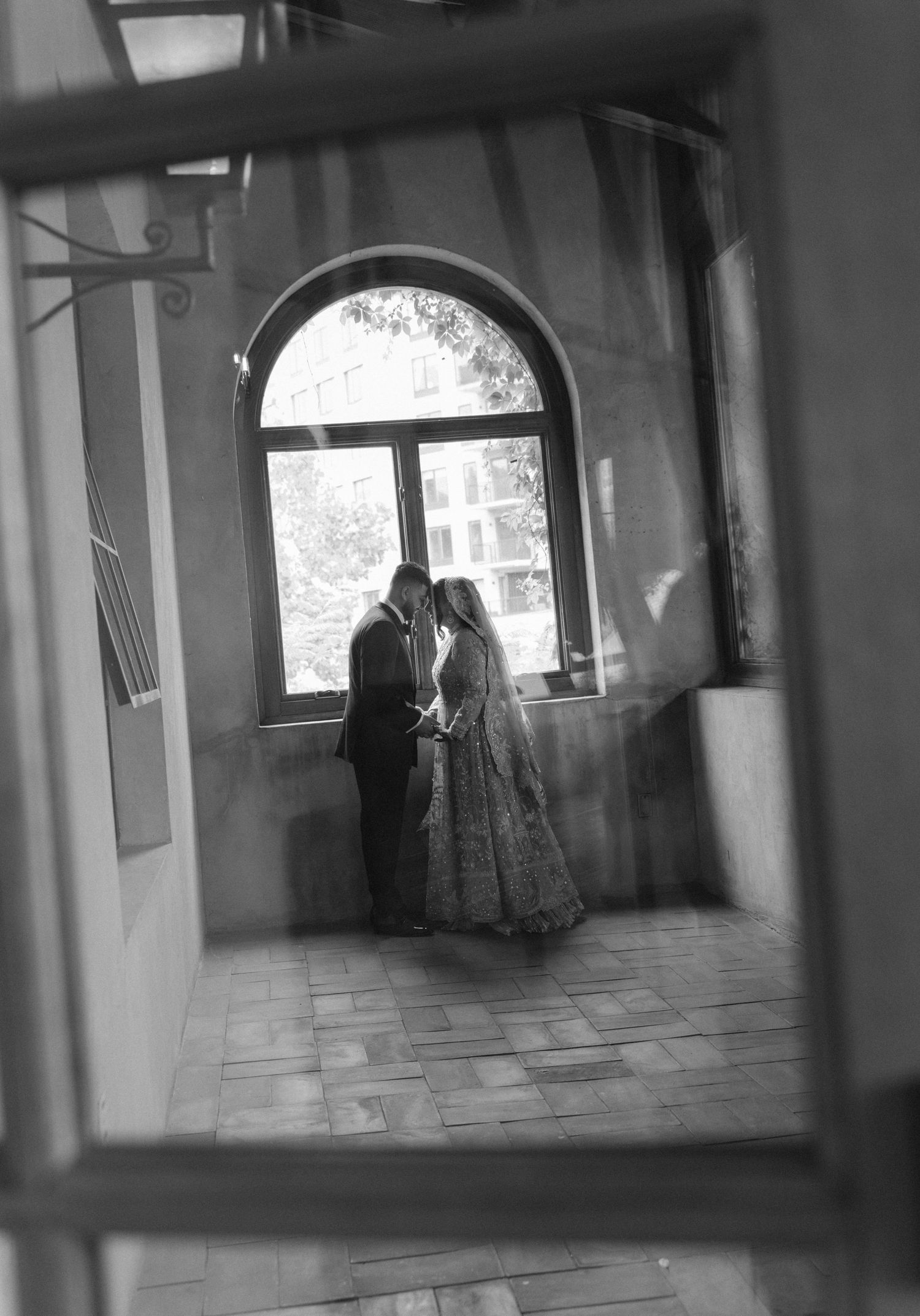Black and white photo of a couple sharing a romantic kiss by an arched window, reflected in a mirror.