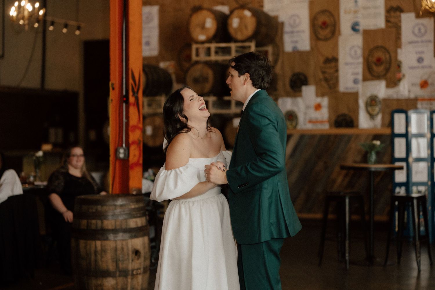Couple shares first dance in rustic brewery wedding venue, bride in white off-shoulder gown, groom in green suit.