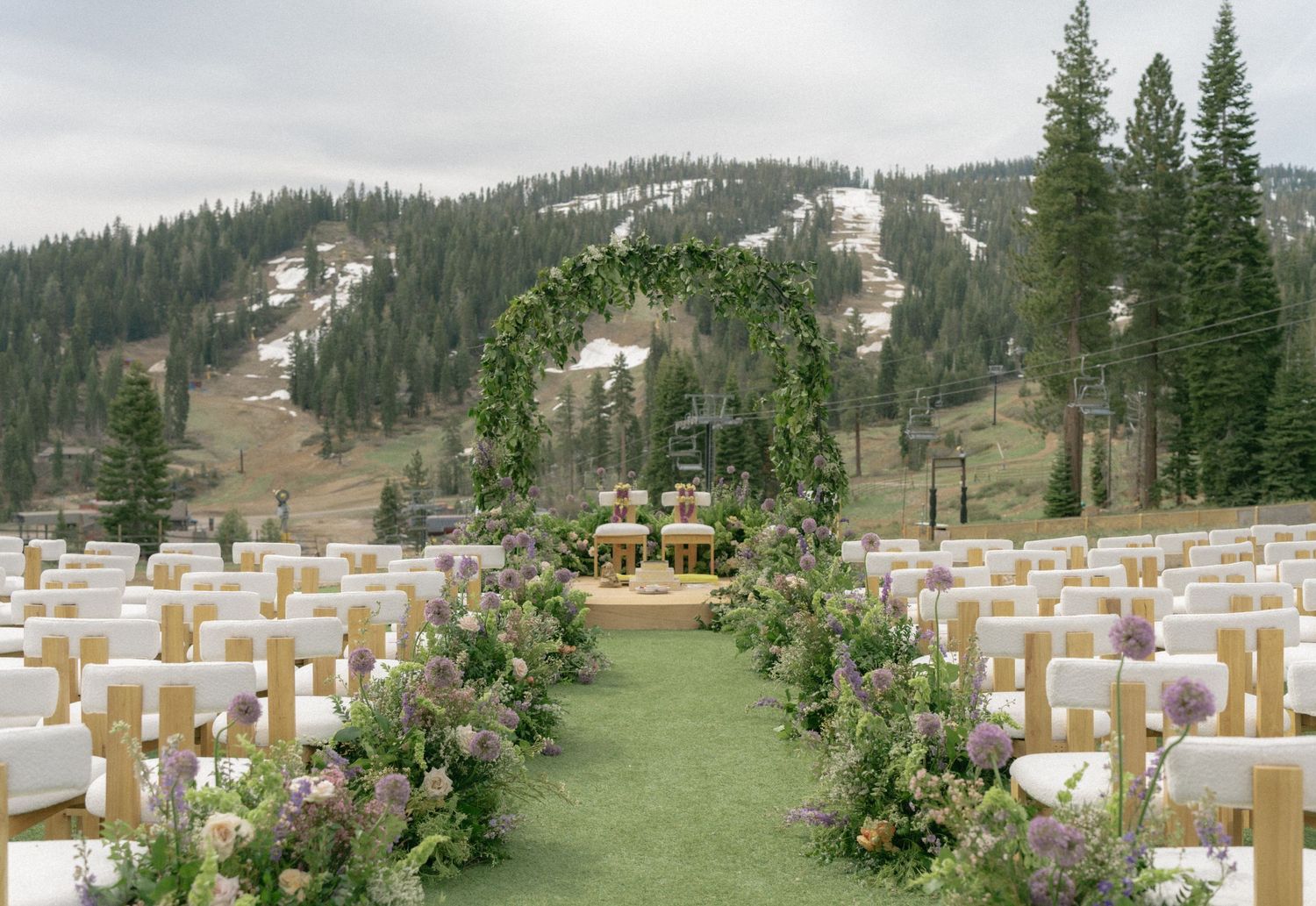 Outdoor mountain wedding ceremony setup with floral arch, white chairs, and pine trees on a scenic slope.