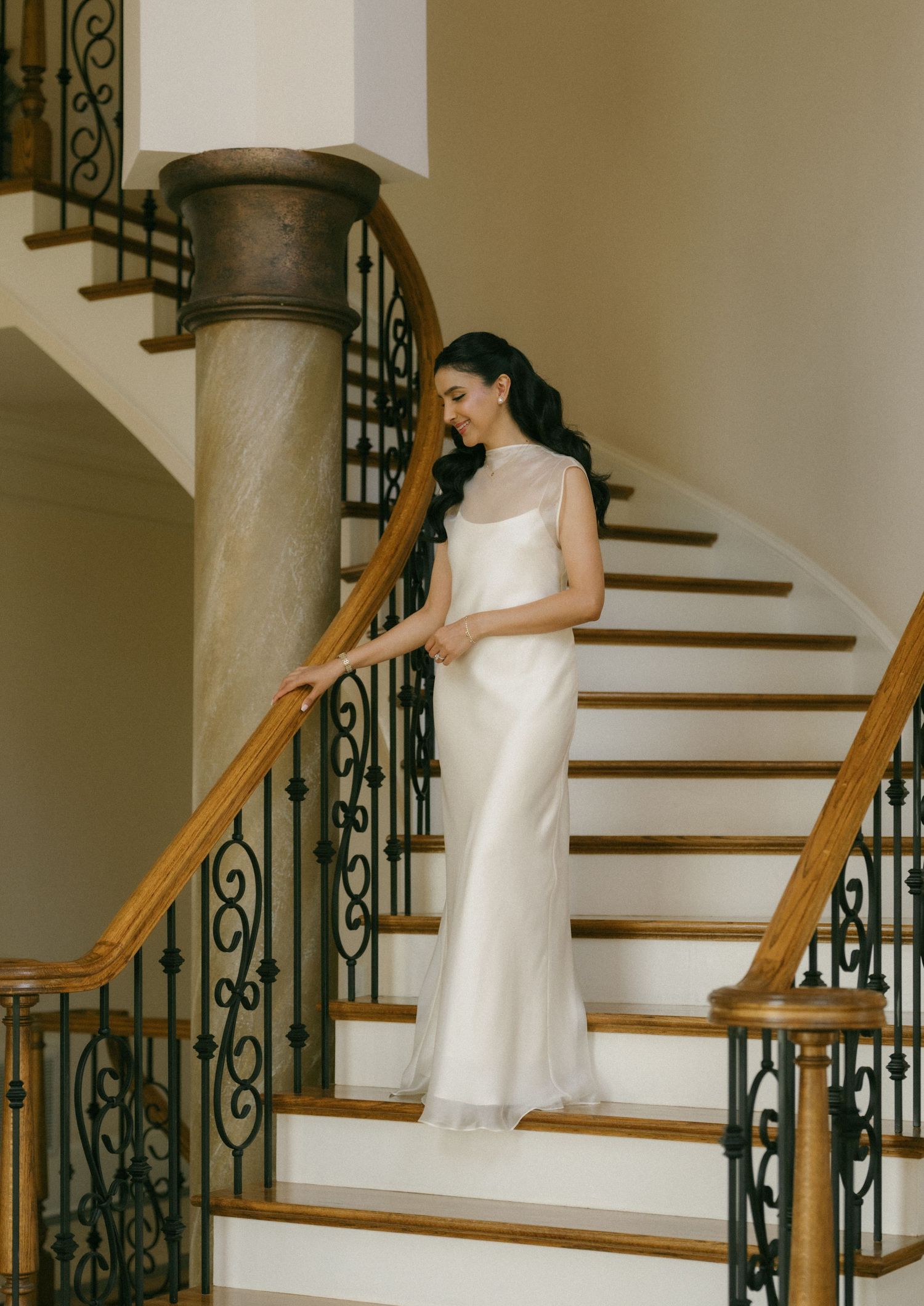 Bride in elegant white satin gown with sheer overlay stands on curved staircase with wrought iron railings.