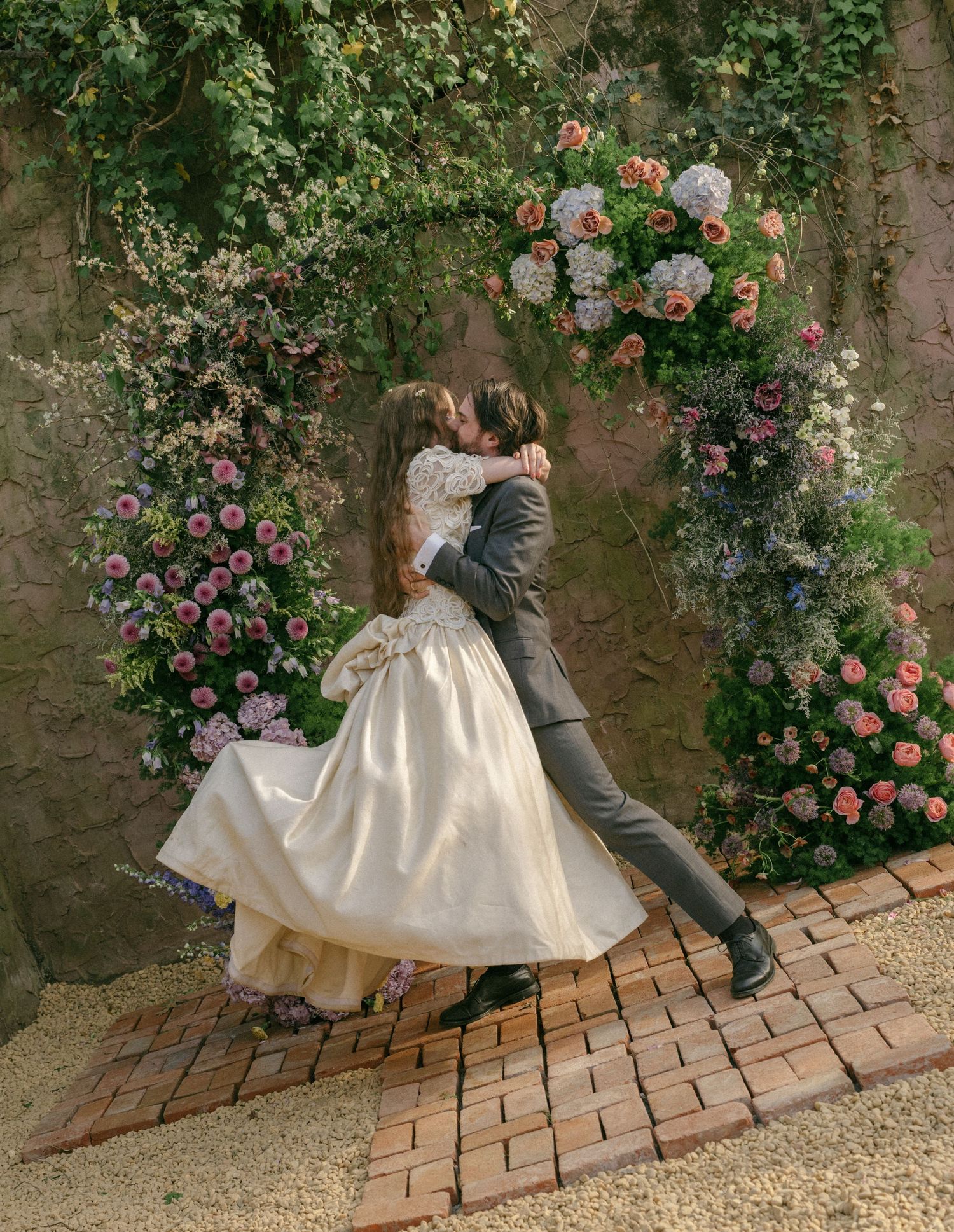 Groom lifts bride for a kiss in front of a lush floral arch with colorful garden blooms.
