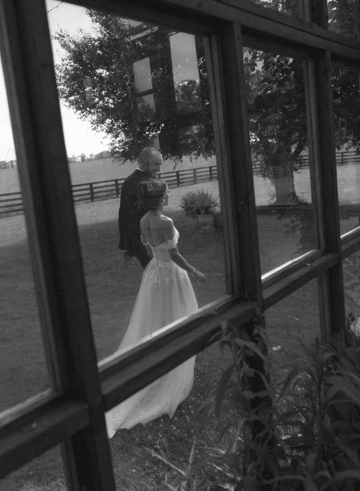 Black and white photo of bride and groom seen through rustic window panes at outdoor farm wedding venue.