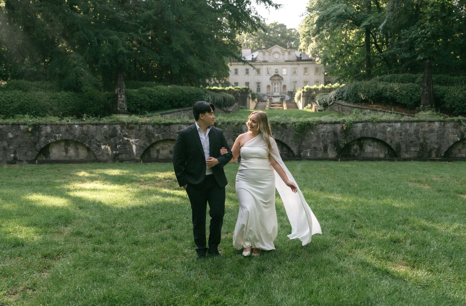 Bride and groom walking hand in hand on lush green lawn with elegant stone mansion and gardens in background.