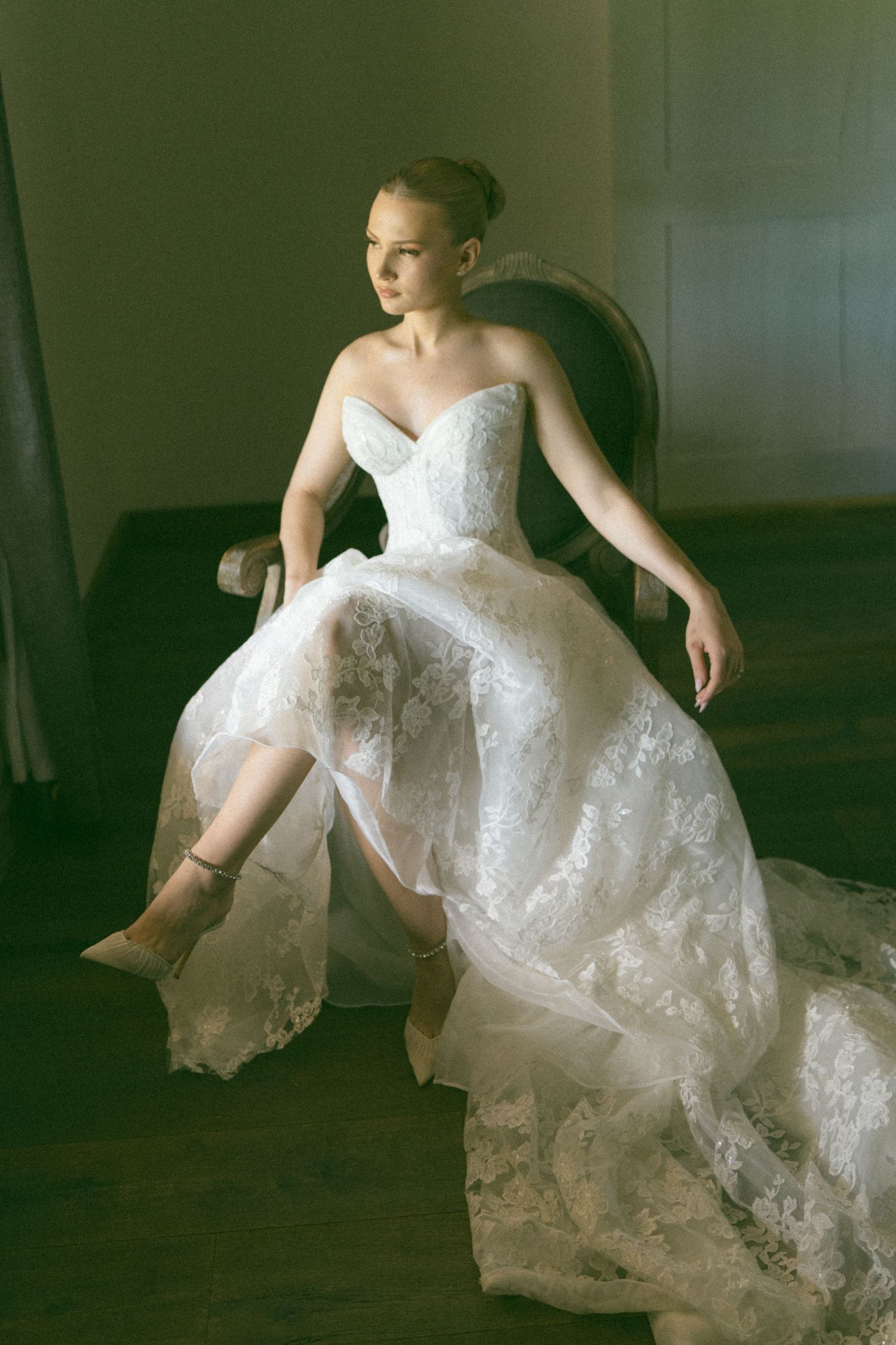Bride in strapless lace ball gown sits gracefully in vintage chair, gazing sideways in soft natural light.
