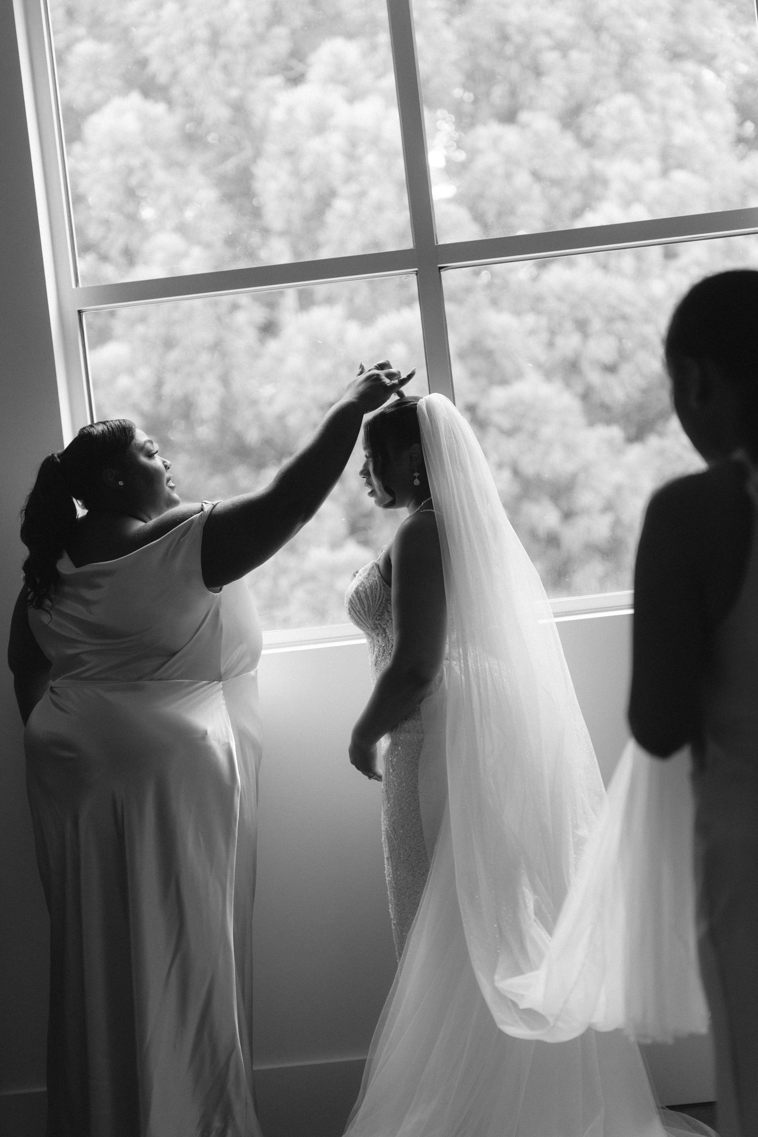 Black and white photo of bridesmaids helping a bride adjust her veil near a large window on her wedding day.