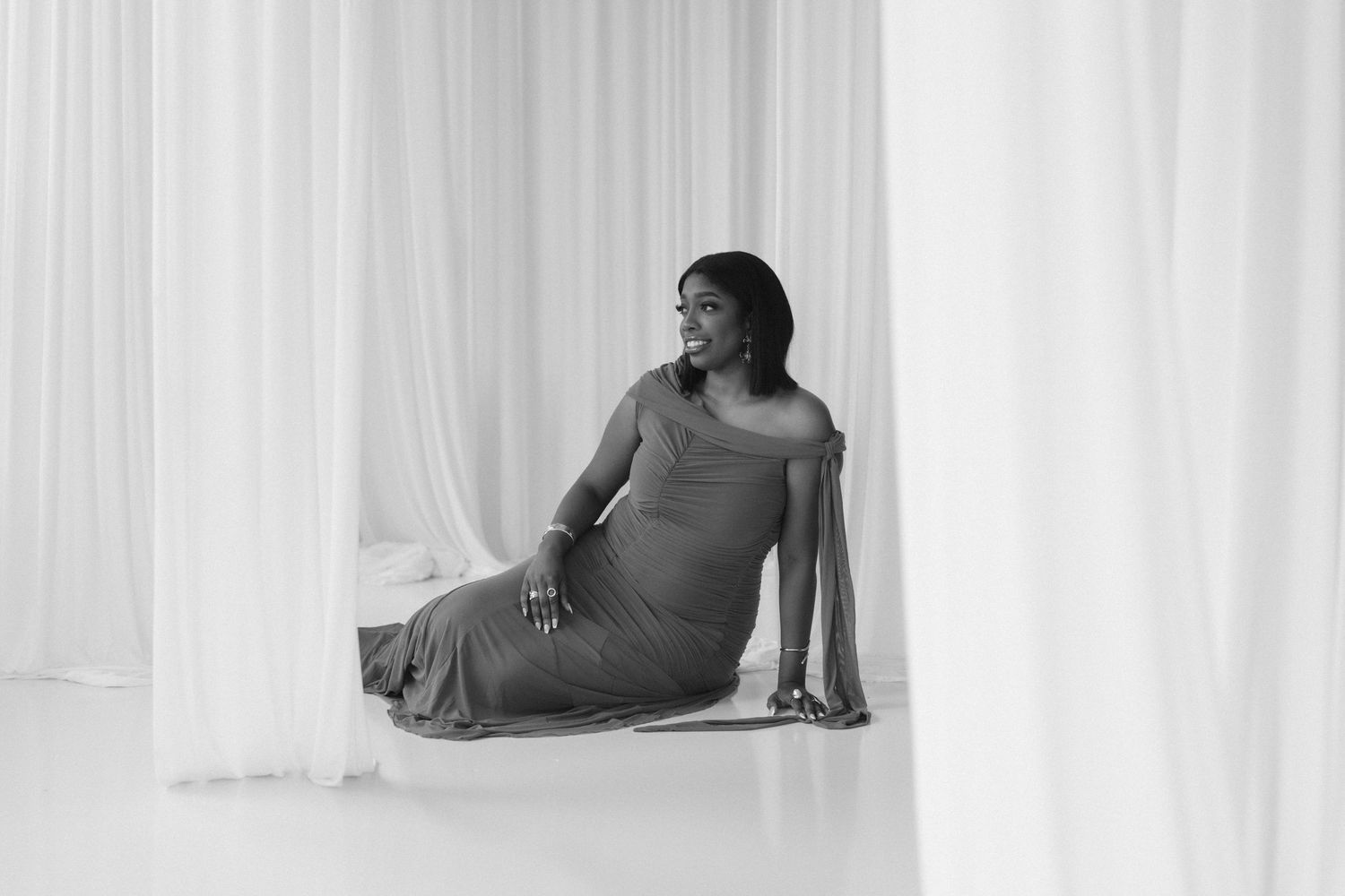 Black and white maternity photo of a woman in a flowing gown sitting gracefully among white curtains.