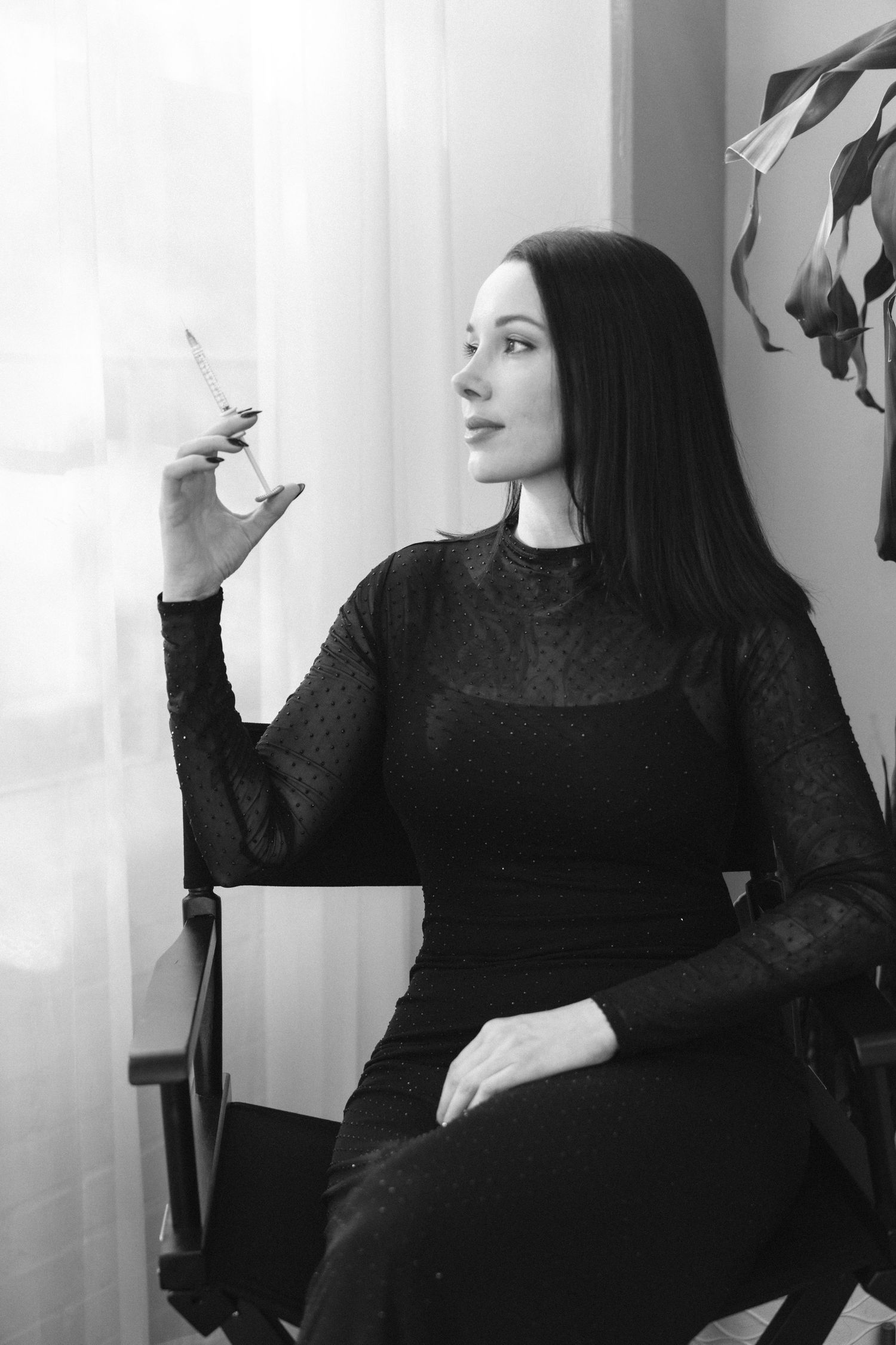 Black and white photo of a woman in a black dress sitting in a chair, holding a wine glass near a window.