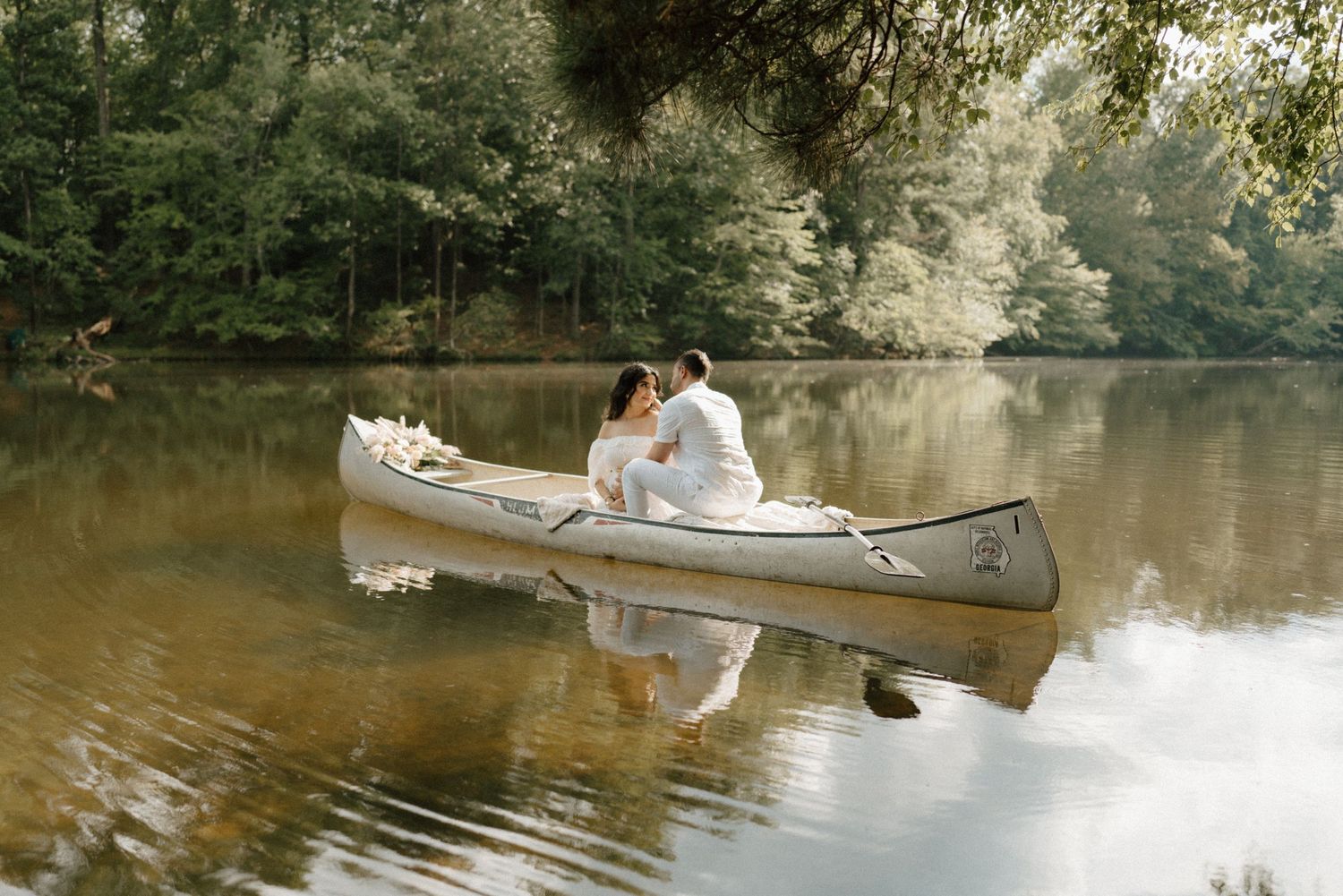 Couple sharing a romantic moment in a white canoe on a serene lake surrounded by lush green trees.