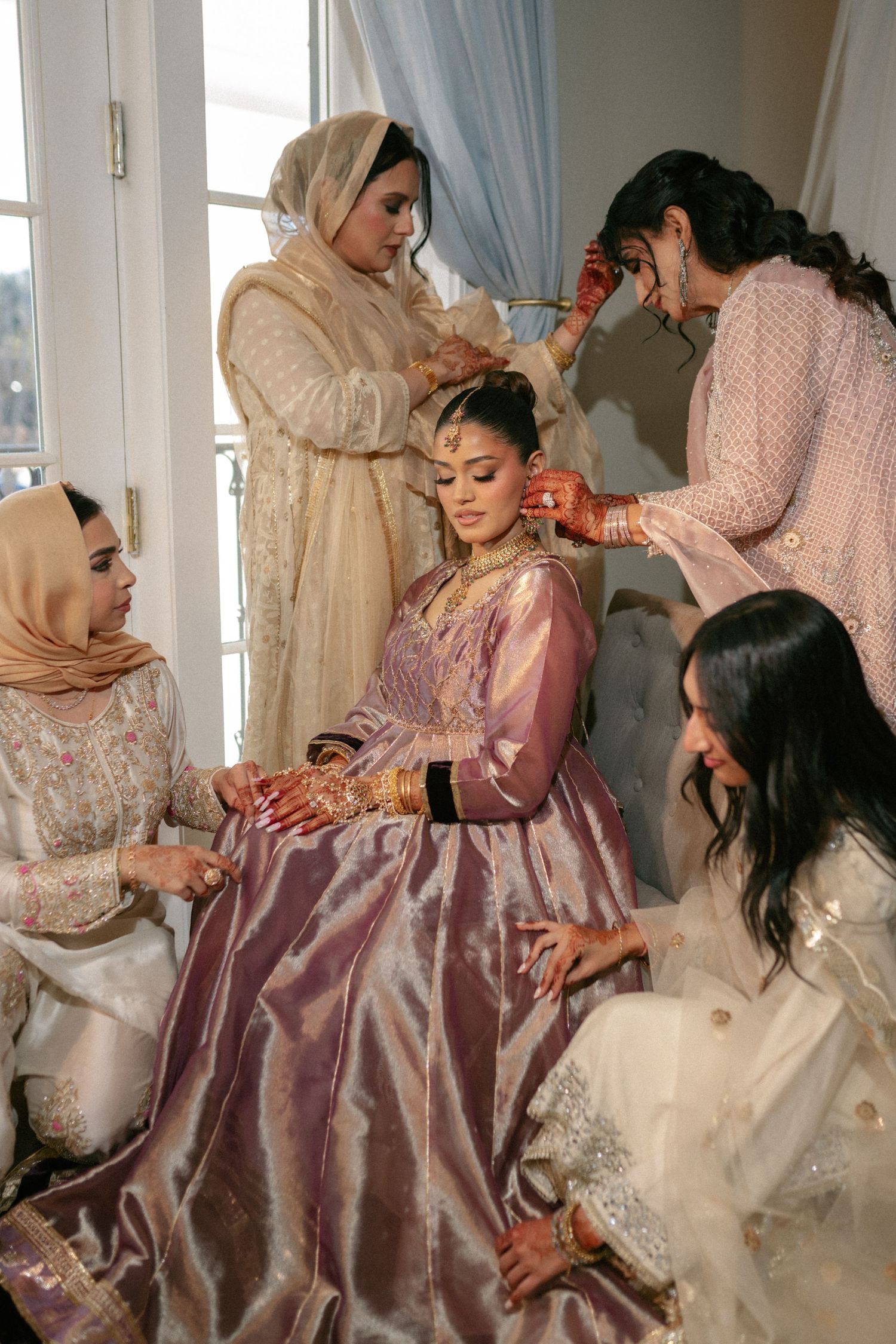 South Asian bride in lavender lehenga being adorned by bridesmaids in white traditional attire before wedding ceremony.