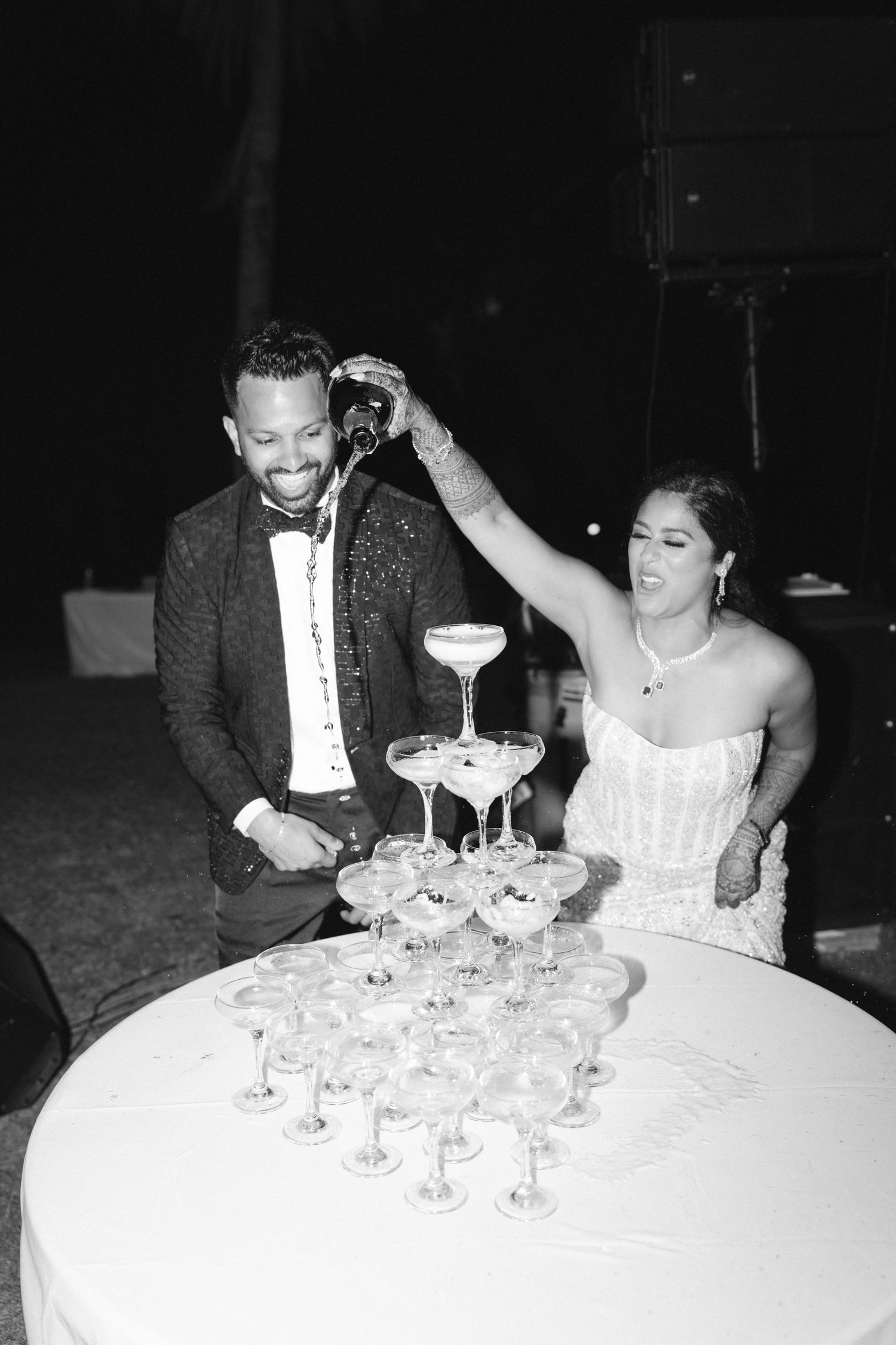 Black and white photo of bride and groom pouring champagne tower at wedding reception celebration.