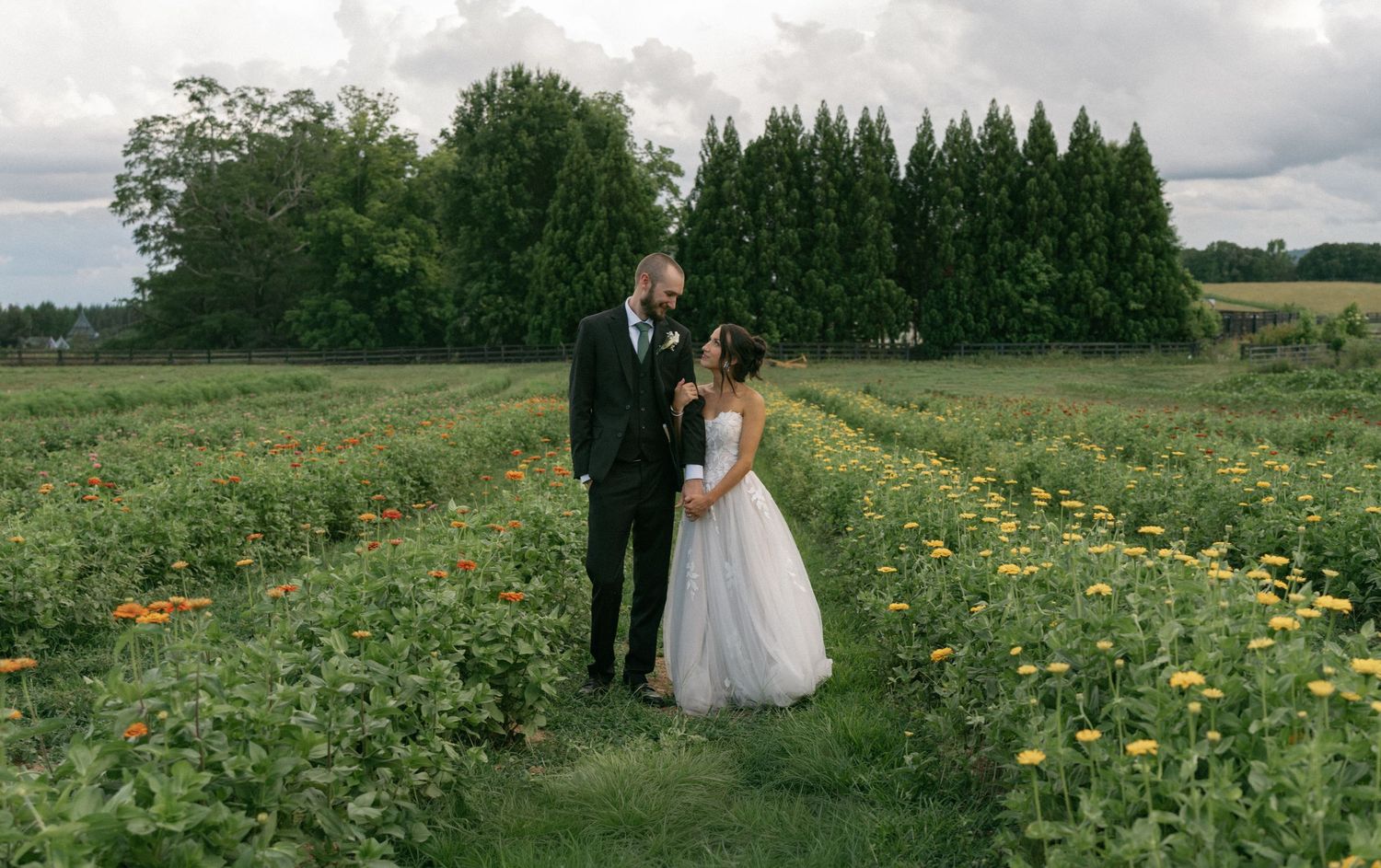 Bride and groom standing in a colorful flower field at an outdoor wedding venue surrounded by lush green trees.
