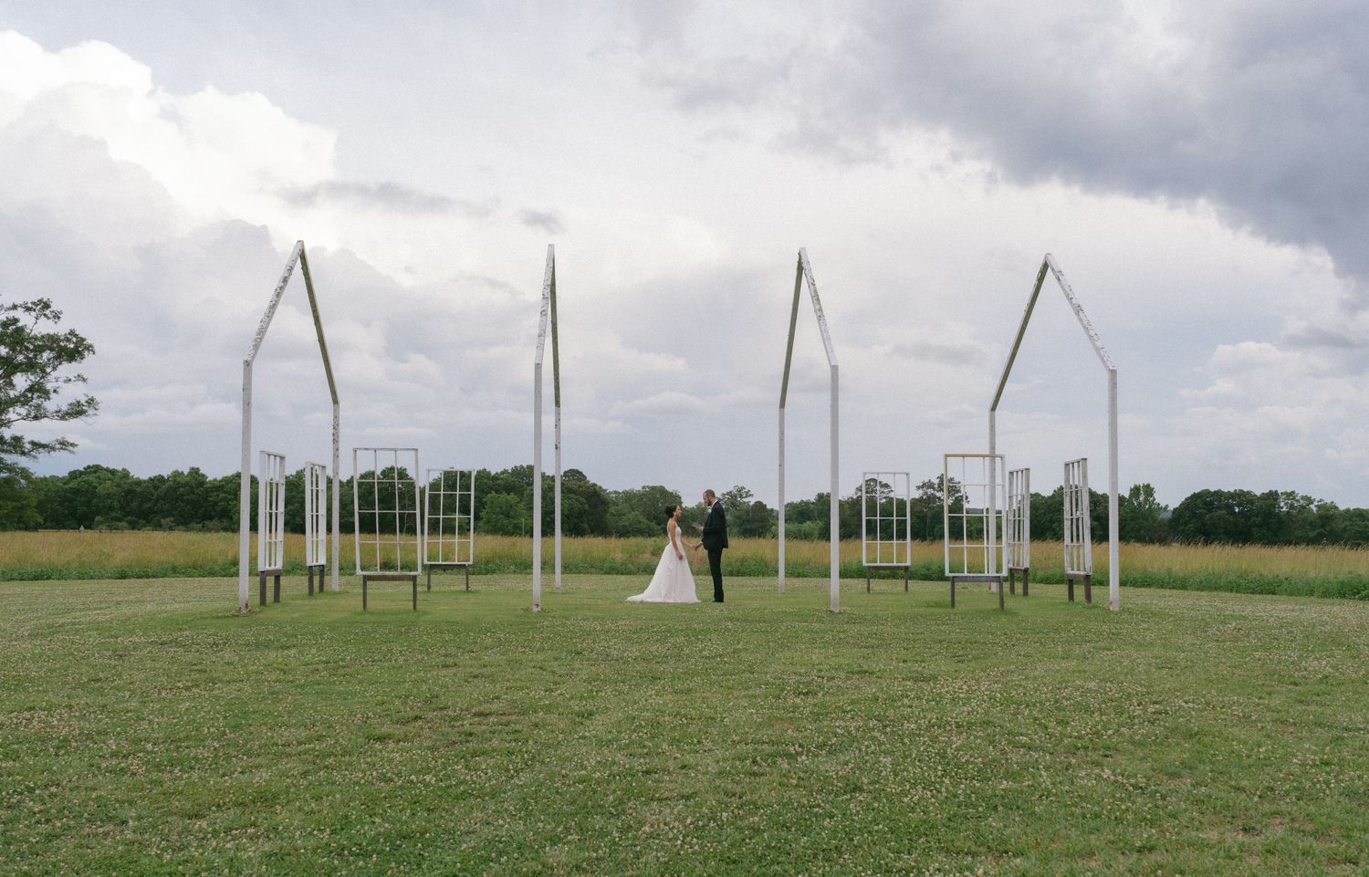 Bride and groom stand among elegant white arch installations in an open green field under a dramatic cloudy sky.