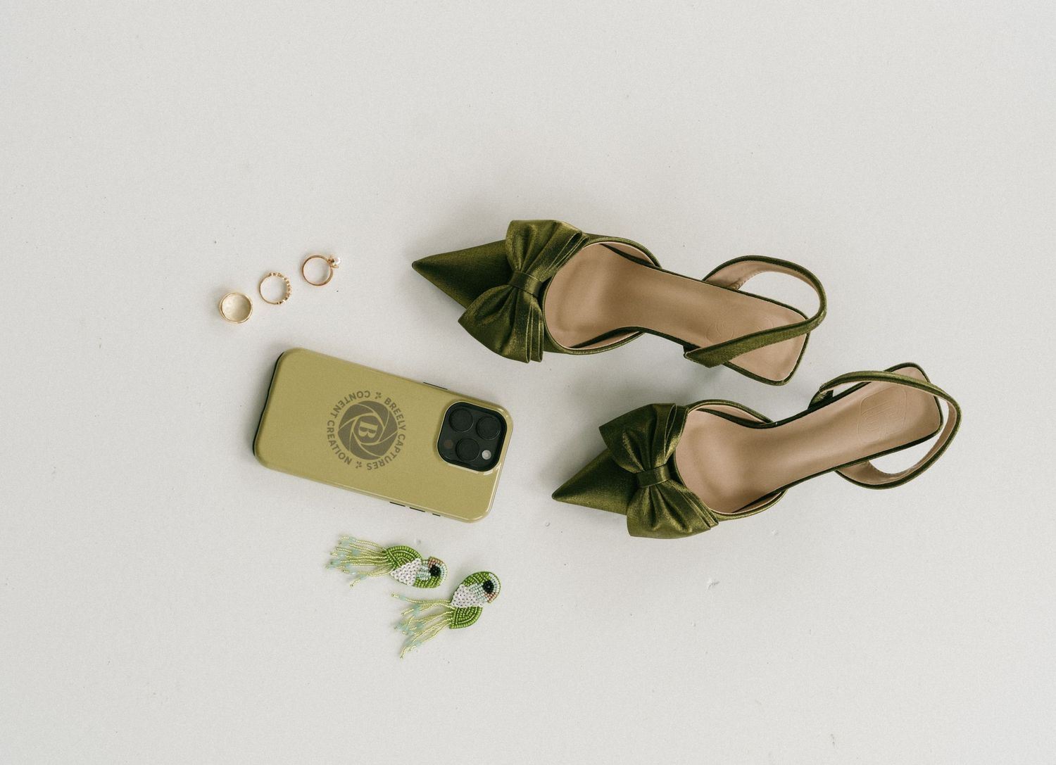 Olive green satin heels with bow detail beside a matching phone case, gold rings, and greenery on white background.