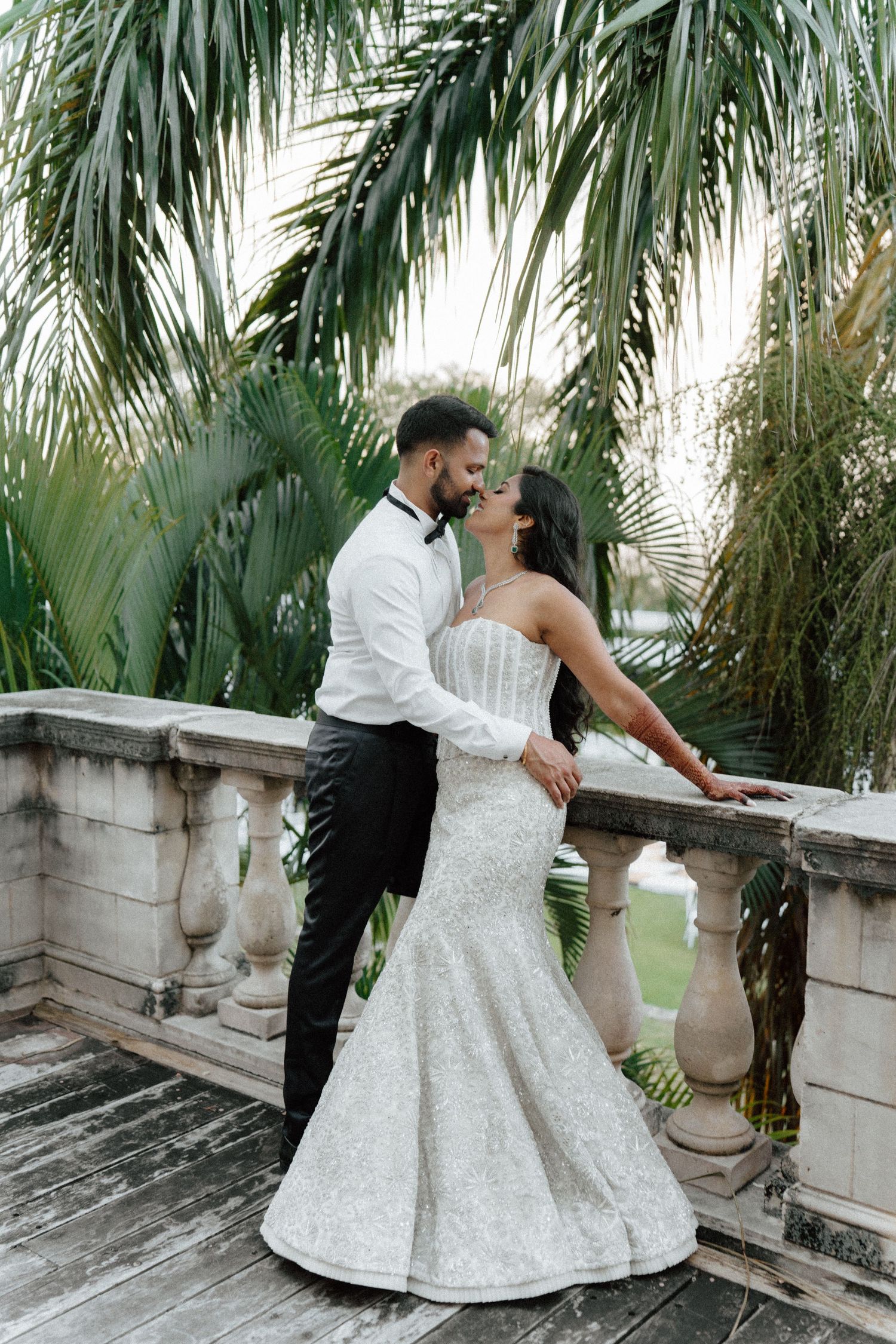 Bride and groom share a romantic moment on a stone balcony surrounded by lush tropical palm trees.