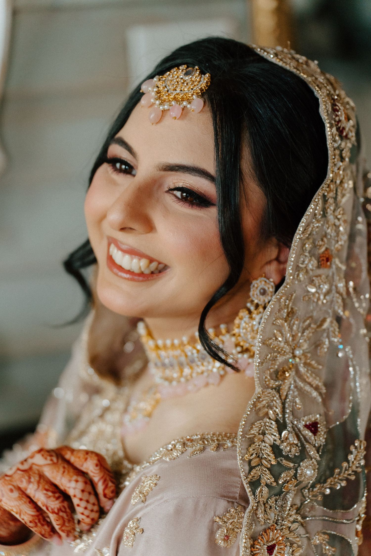 Smiling South Asian bride wearing gold maang tikka, embroidered dupatta, layered necklace, and mehndi on her hands.