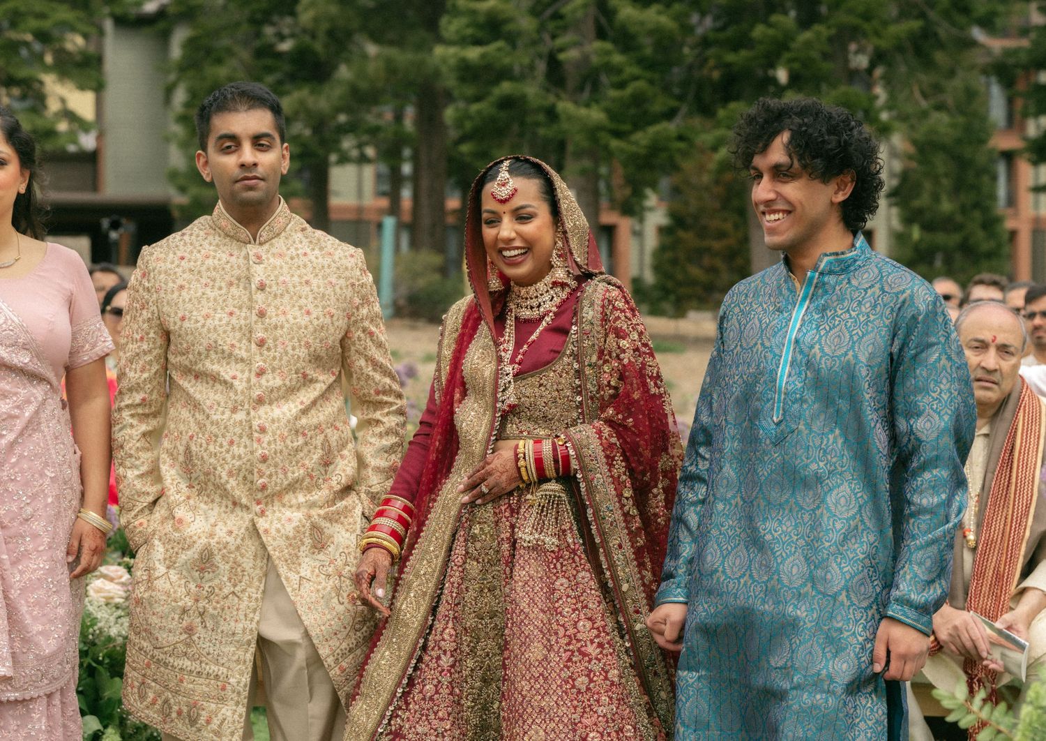 Bride in red lehenga walks with groom in gold sherwani at Indian wedding ceremony outdoors.