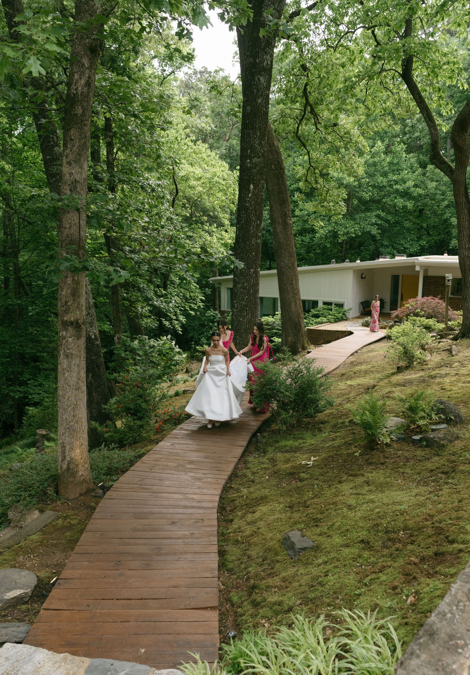 Bride and bridesmaids in pink dresses walking along wooden boardwalk path through lush green forest to venue.