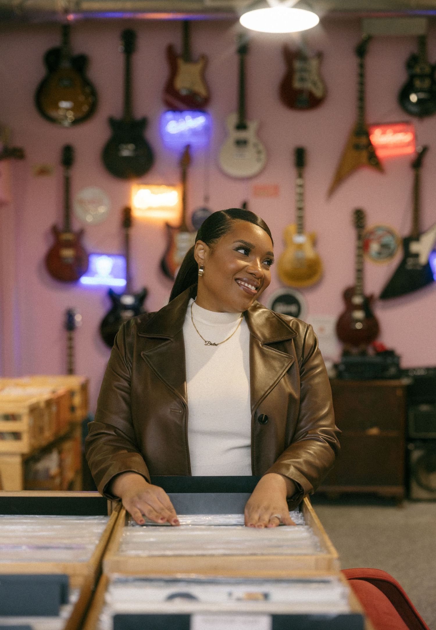 A smiling woman in a brown leather jacket browses vinyl records in a music store with guitars displayed on a pink wall.