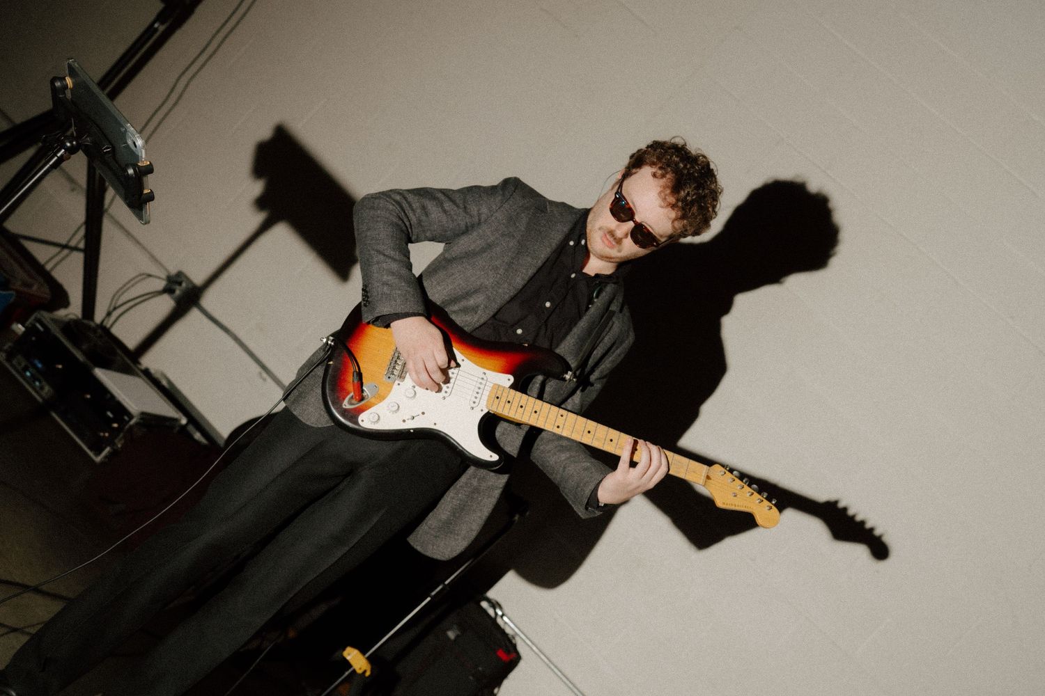 Musician playing a sunburst Fender Stratocaster guitar on stage wearing sunglasses and a gray jacket.