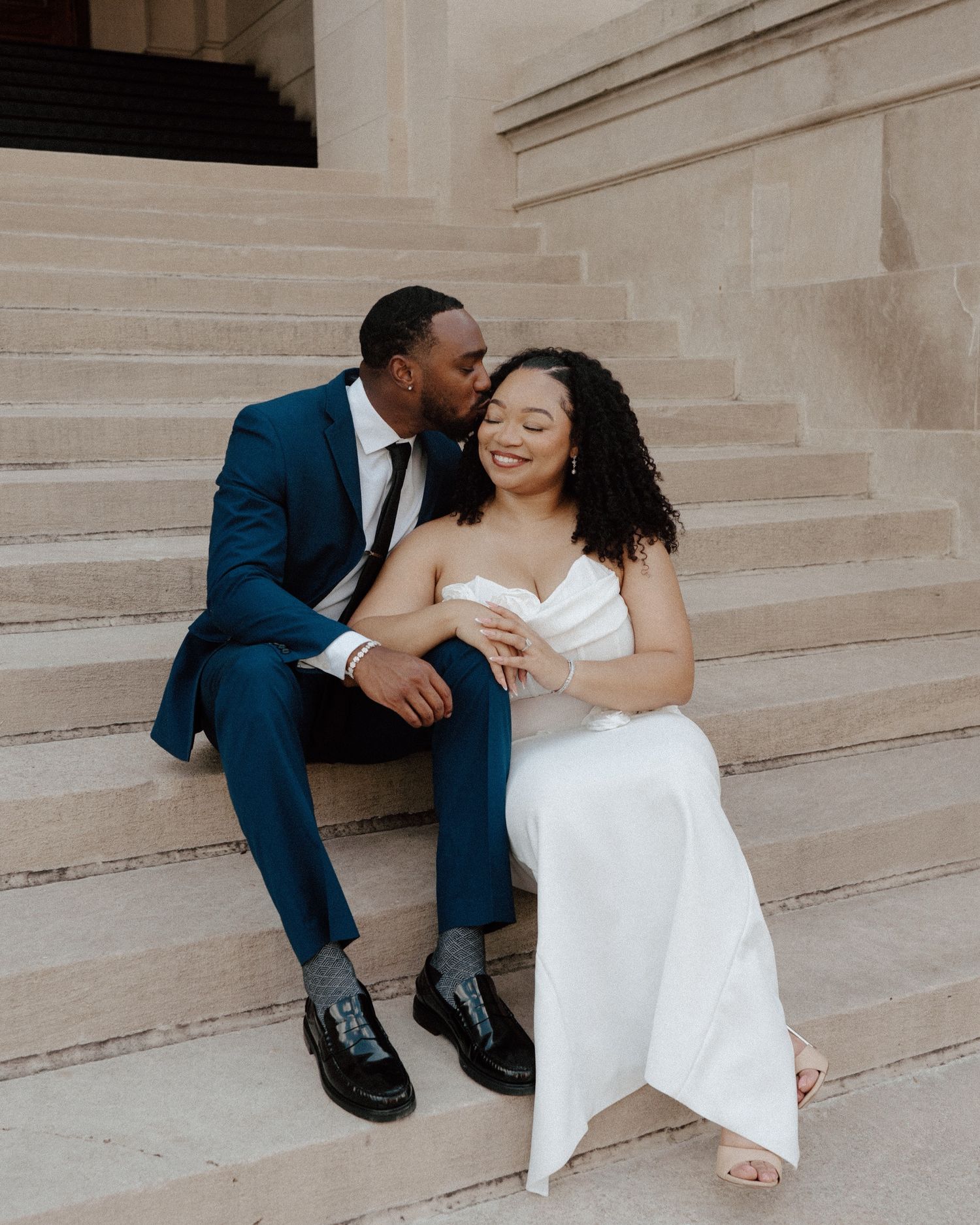 A couple in wedding attire sits on stone steps, the groom in a blue suit kissing the smiling bride in a white gown.