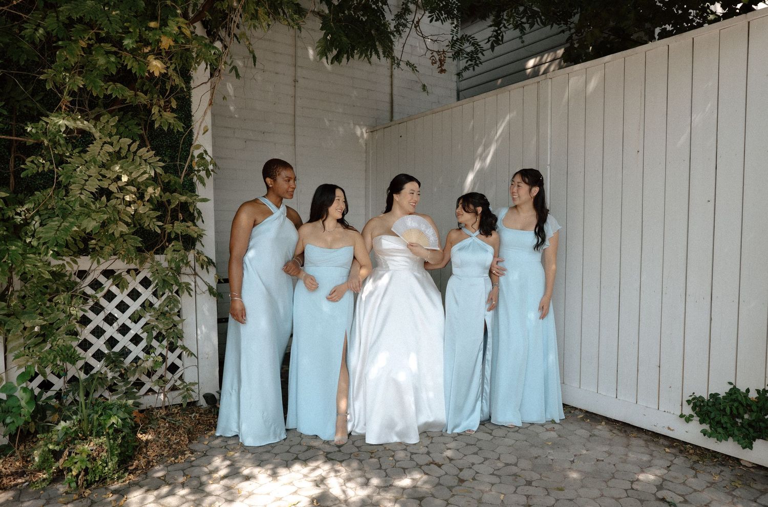 Bride in white gown with four bridesmaids in light blue dresses posing outdoors near a white fence.