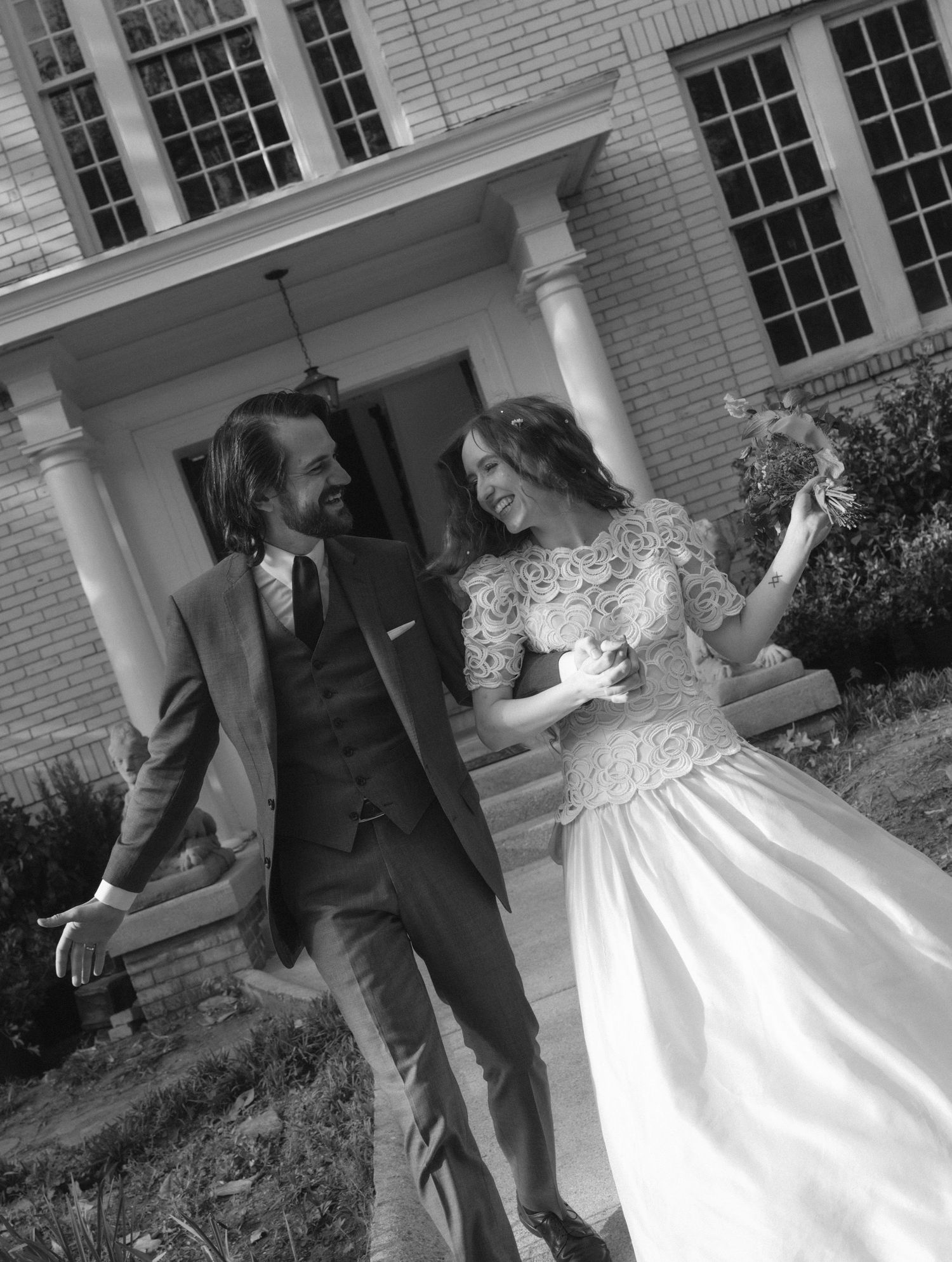 Black and white photo of a laughing bride and groom walking together in front of a classic colonial building.
