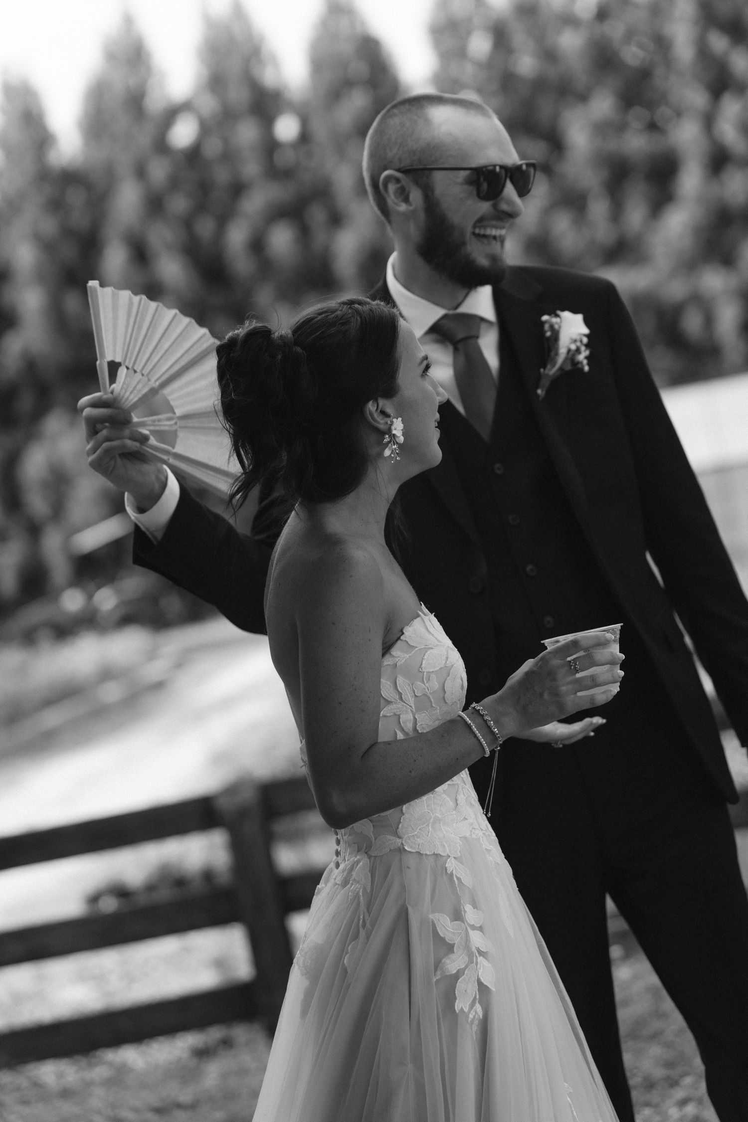 Black and white photo of a smiling bride holding a fan and groom in sunglasses at an outdoor wedding.