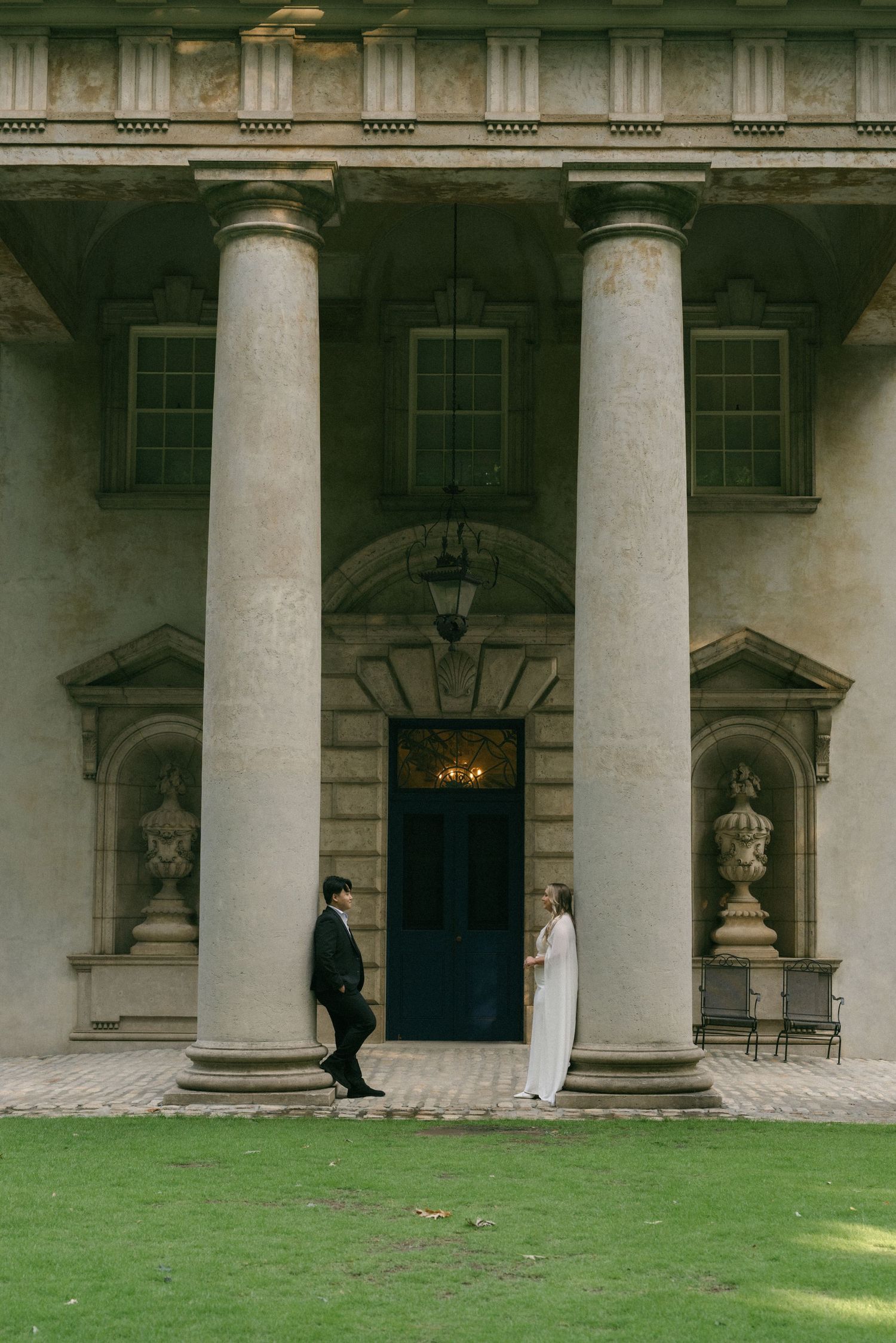 Couple in wedding attire standing between grand stone columns at a classical Georgian mansion entrance.