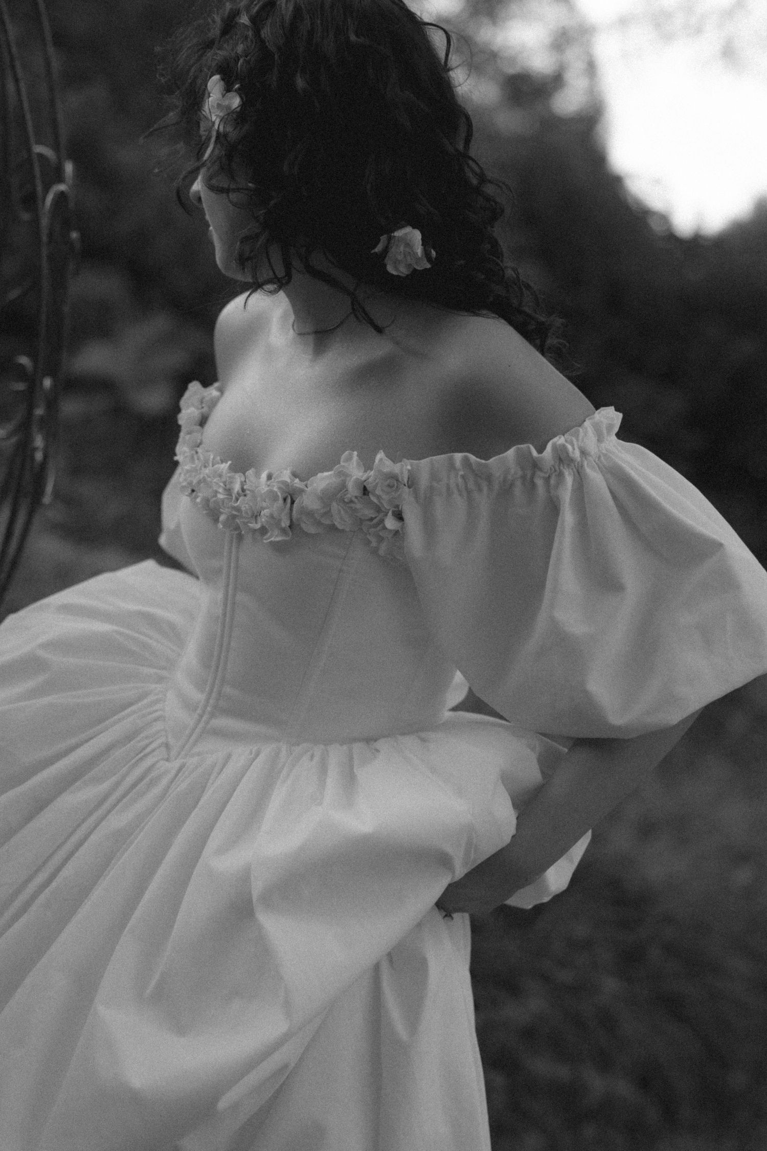 Black and white photo of a woman in an off-shoulder white ballgown with puff sleeves and floral details in her curly hair.