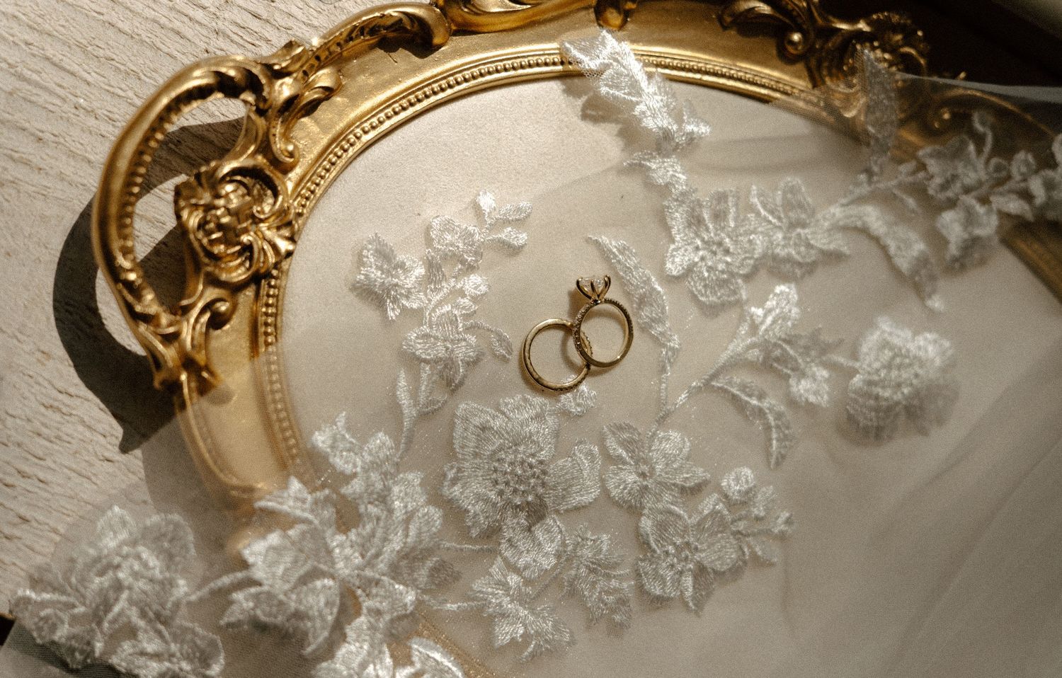 Gold ornate mirror frame with white lace floral fabric and two wedding rings displayed elegantly.