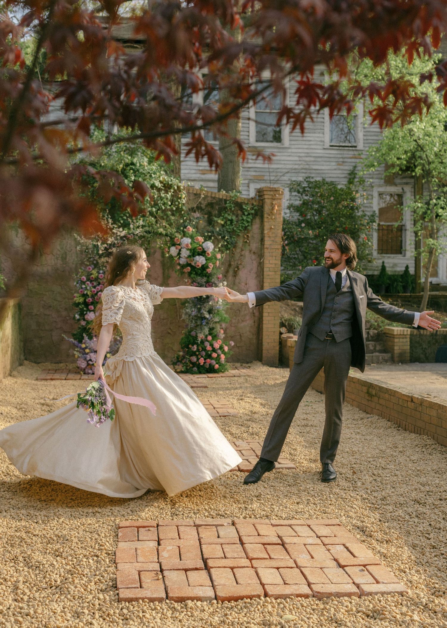 Bride and groom dancing joyfully in a sunlit garden with a floral arch and red maple tree backdrop.