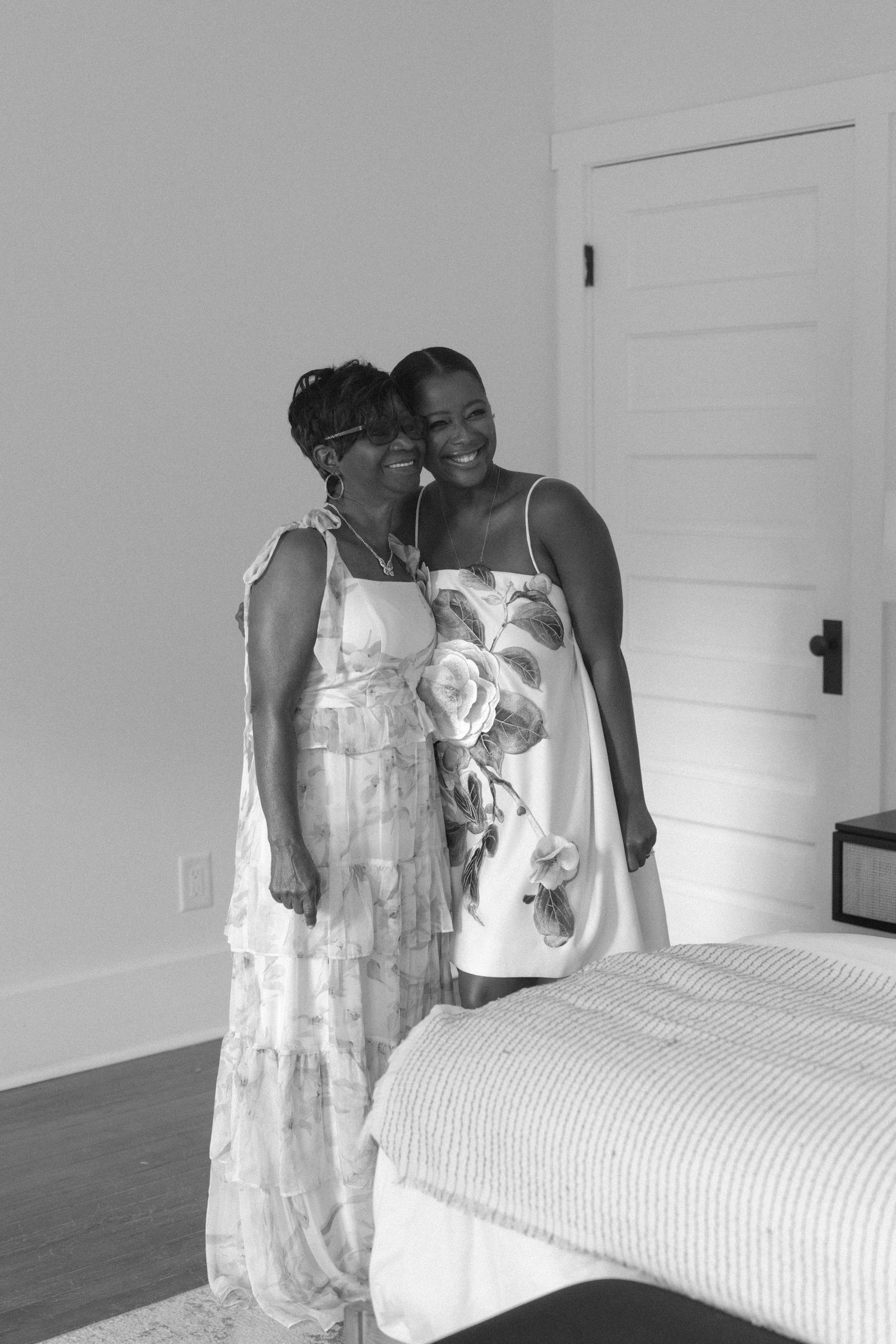 Two women in summer dresses smiling together in a bright bedroom, black and white photo.