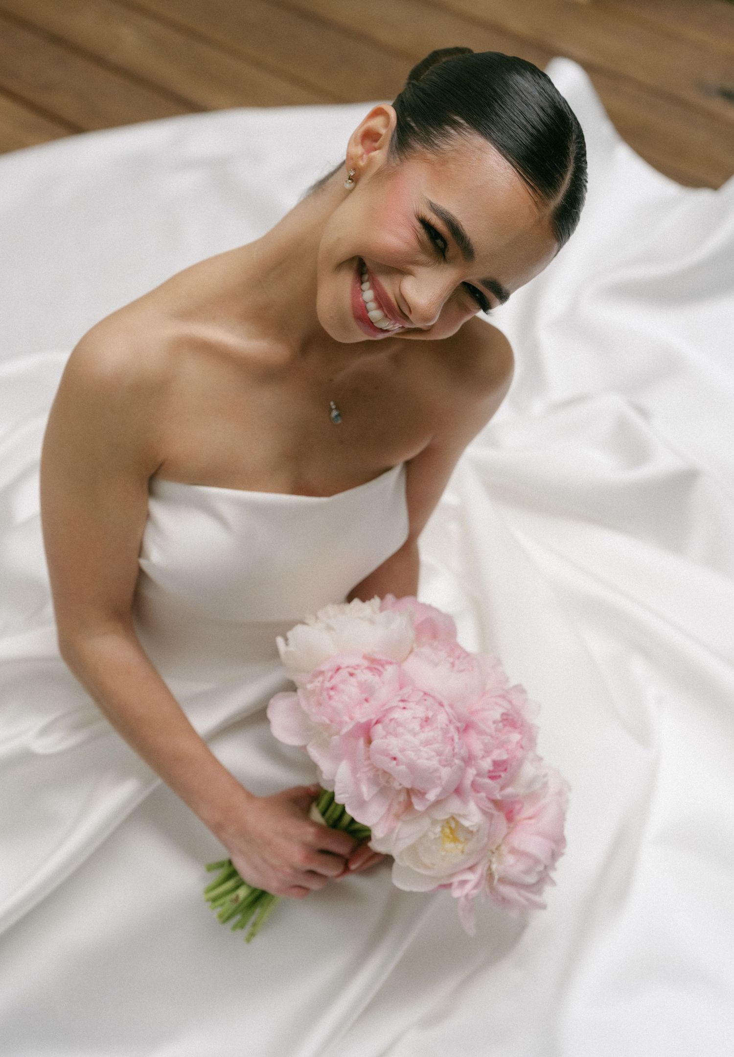 Smiling bride in strapless white gown holding pink peony bouquet while seated on flowing dress train.