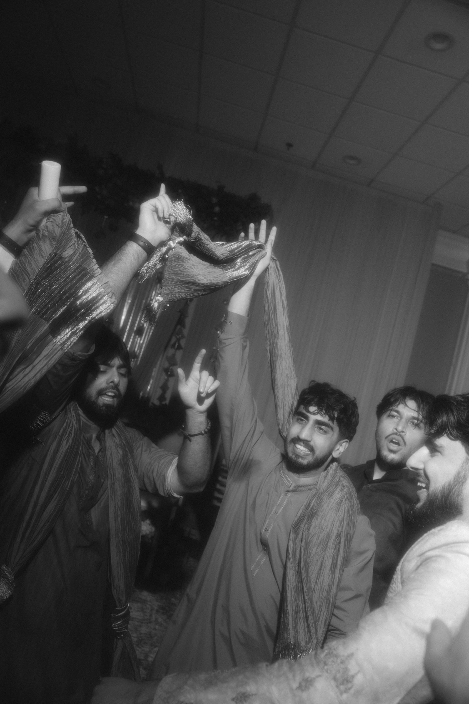 Black and white photo of young men celebrating at a party, raising hands and cheering in traditional South Asian attire.