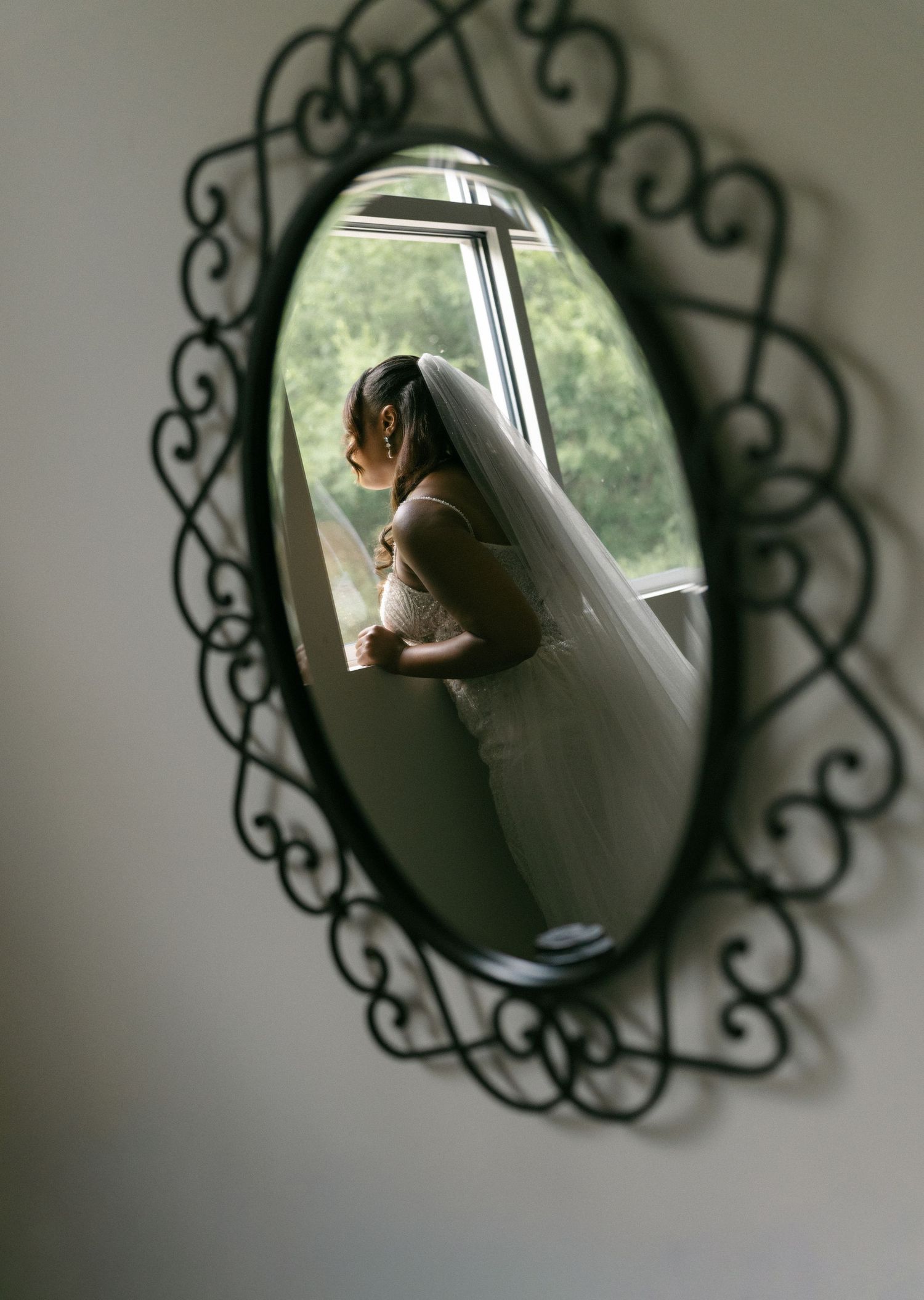 Bride in a white gown and veil reflected in an ornate oval mirror by a window.