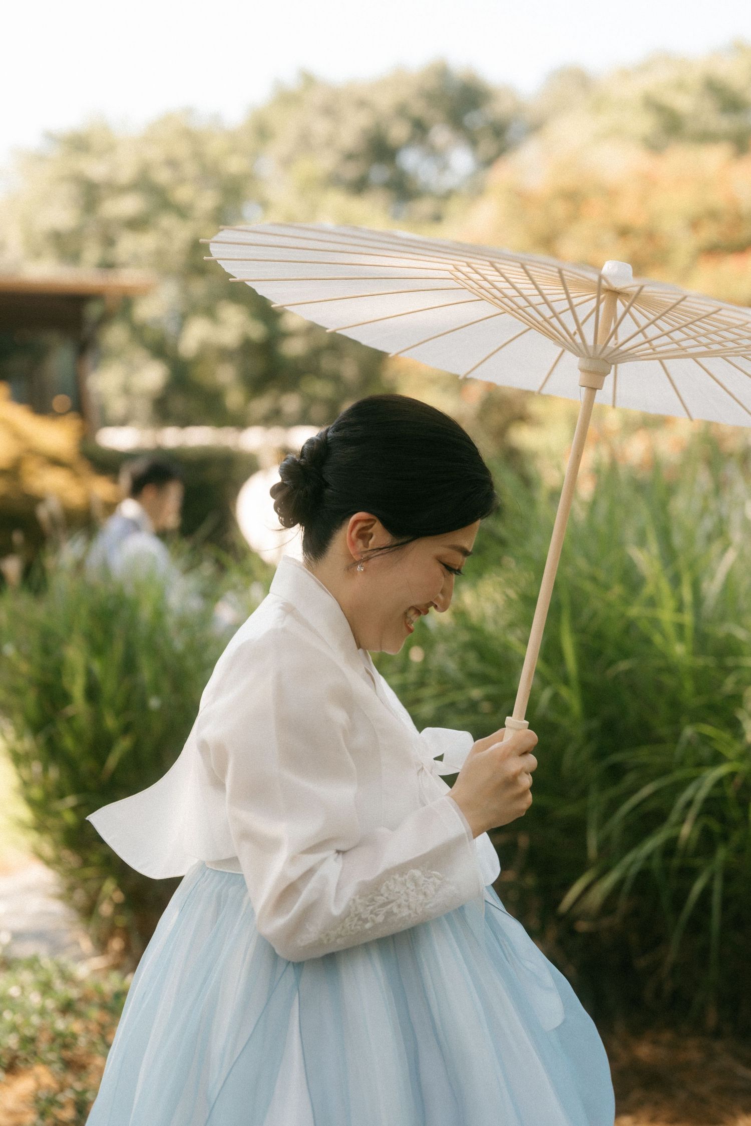 Woman in traditional Korean hanbok with white top and blue skirt holds paper parasol outdoors.