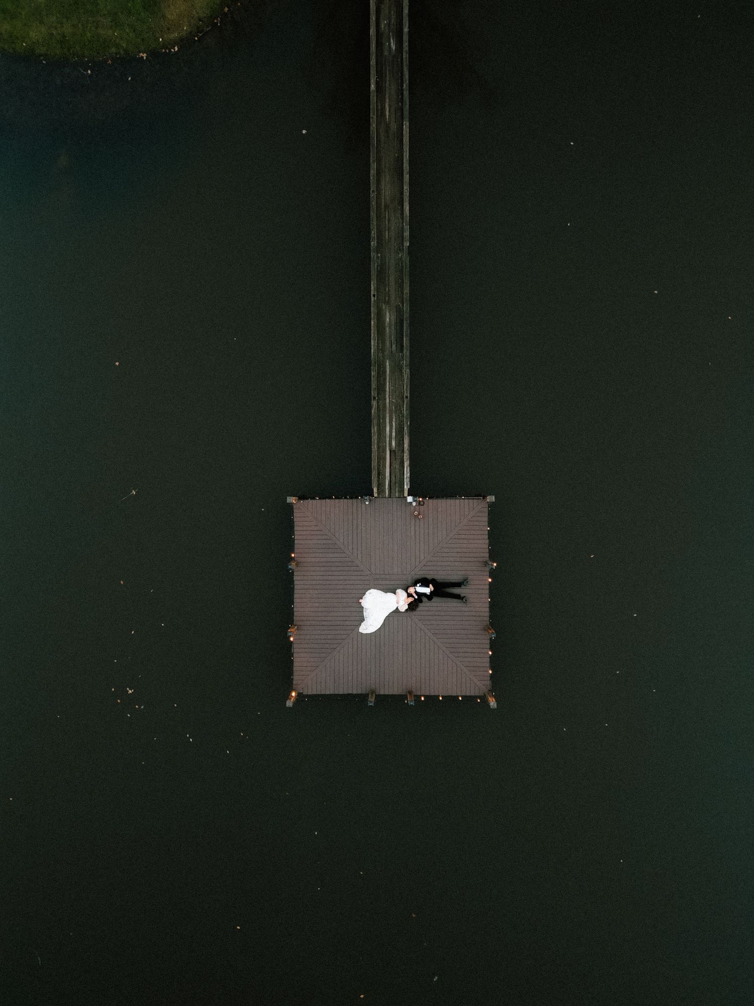 Aerial drone view of a bride and groom lying on a wooden dock platform surrounded by dark green water.