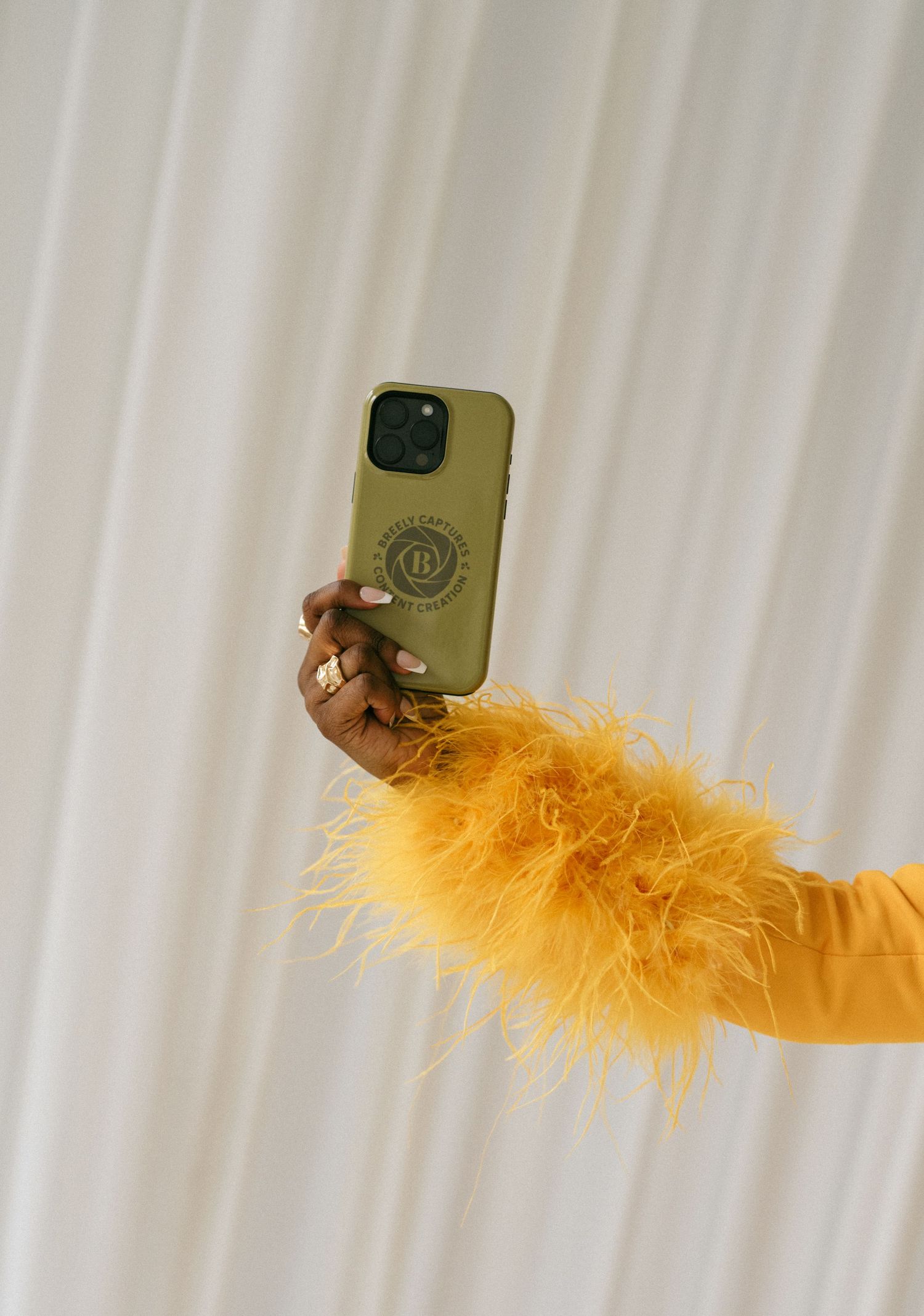 A hand with yellow feather sleeve holds an olive green iPhone case with a logo against a white curtain backdrop.
