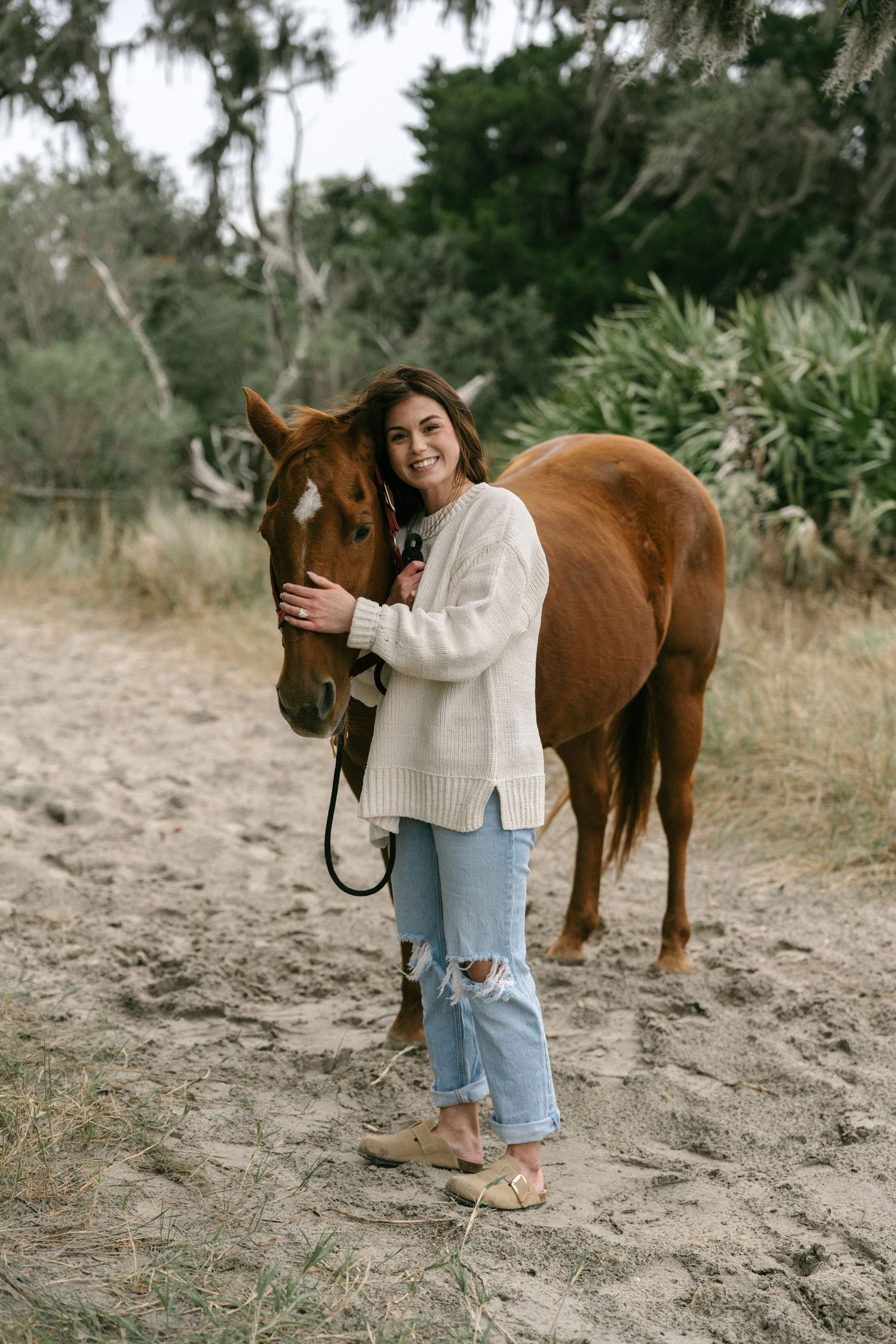 Young woman in cream sweater and ripped jeans smiling while hugging a chestnut horse on a sandy path.