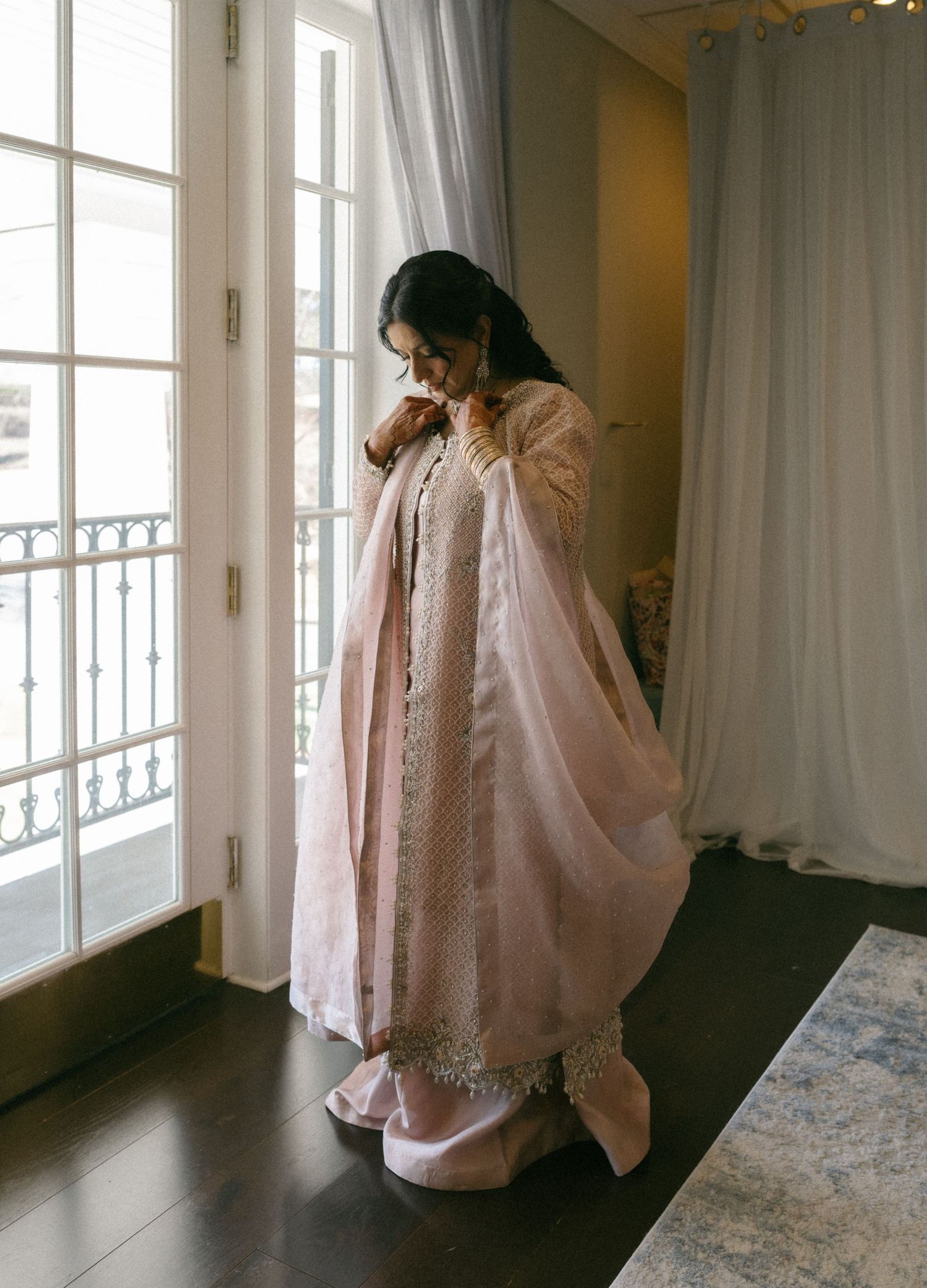 Woman in elegant blush pink Indian anarkali suit with dupatta stands by French doors in a softly lit room.