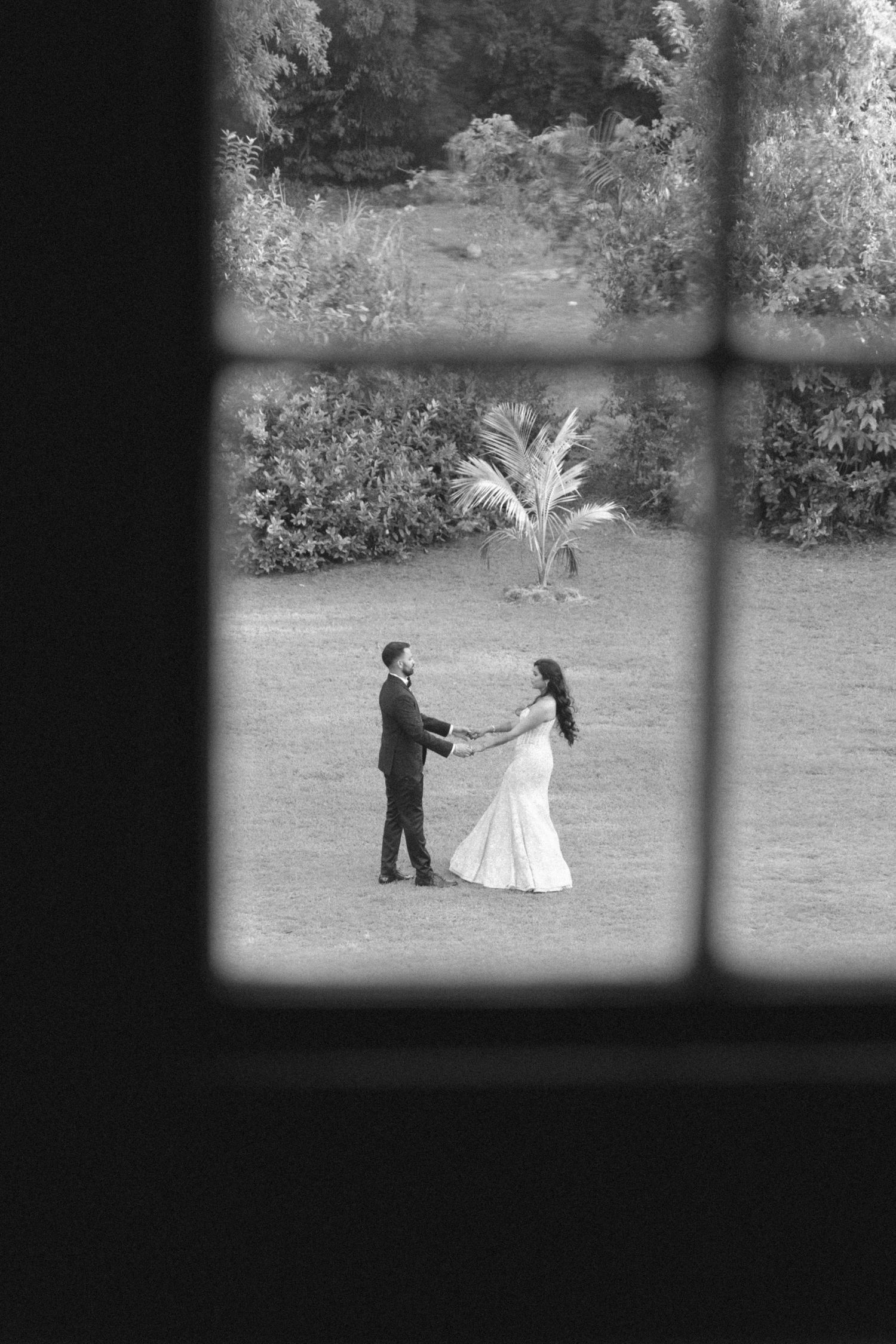 Black and white photo of bride and groom holding hands in lush garden, viewed through window frame.