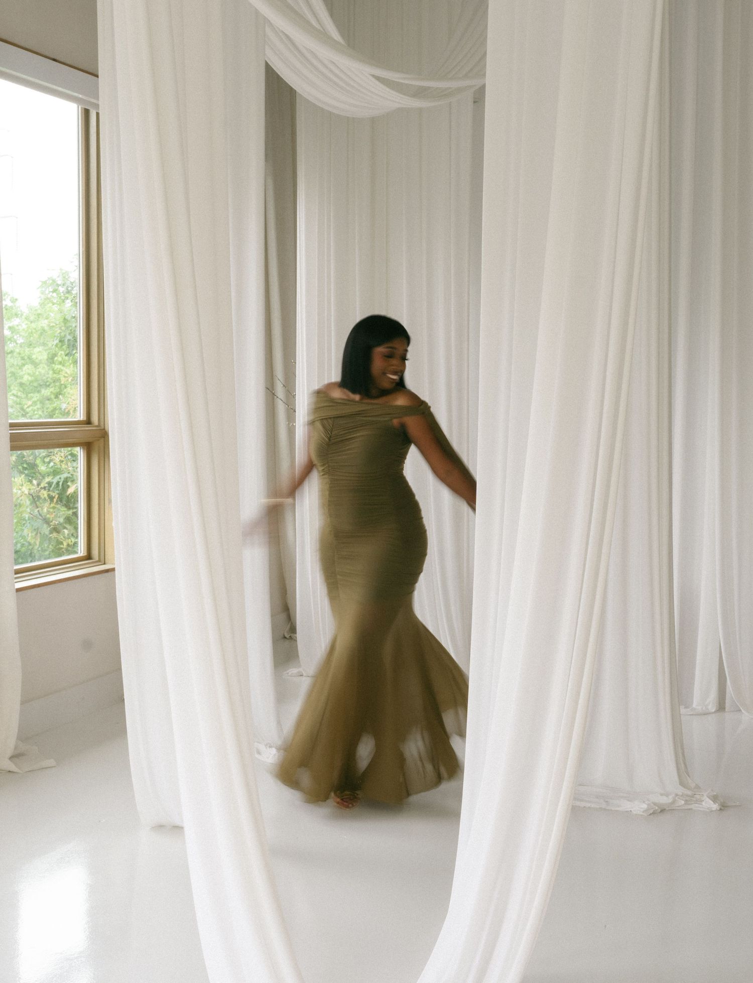 Woman in olive green mermaid gown dancing gracefully among flowing white curtains in an elegant bright studio.