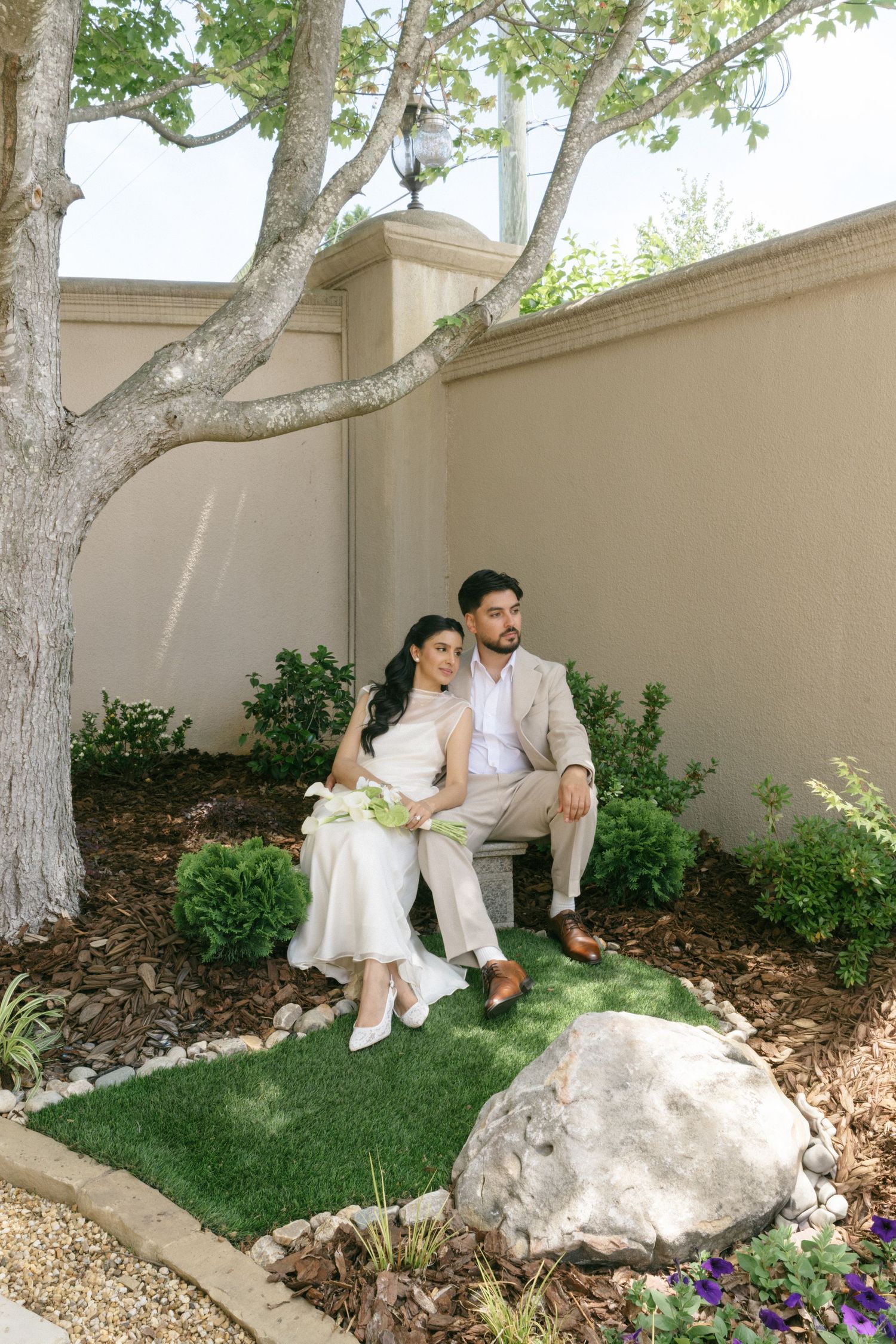 A bride and groom sit together on a bench in a garden courtyard, surrounded by greenery and a large tree.